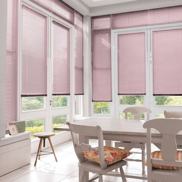 Pink horizontal Venetian blinds covering bay windows, lowered and filtering daylight (some panels slightly raised at bottoms), in a bright dining nook with white table and chairs and a garden view.