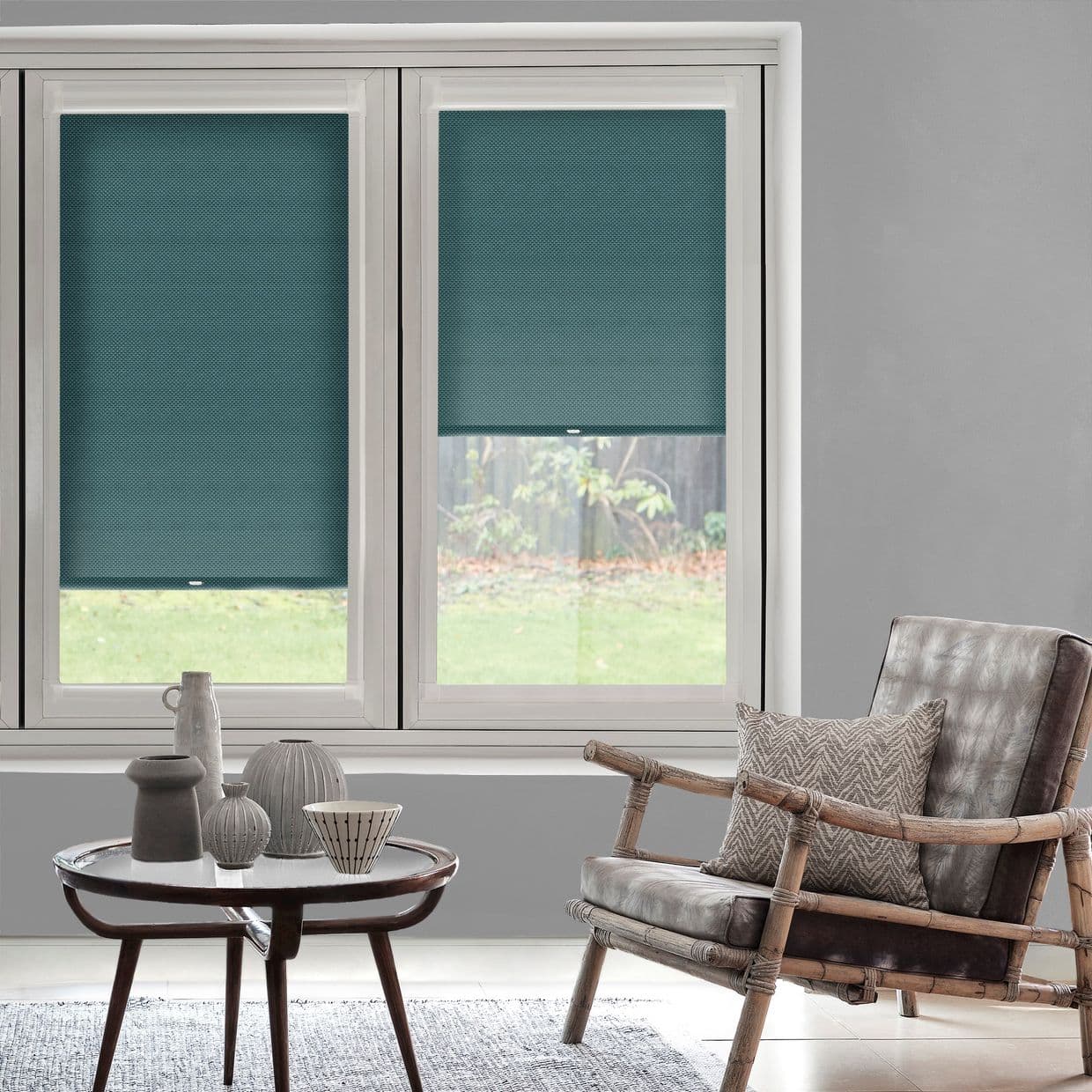 Teal cellular roller blinds, lowered across two adjacent windows, partially blocking view and filtering daylight; set in a gray-walled living room with rattan armchair, cushion, round coffee table and vases.
