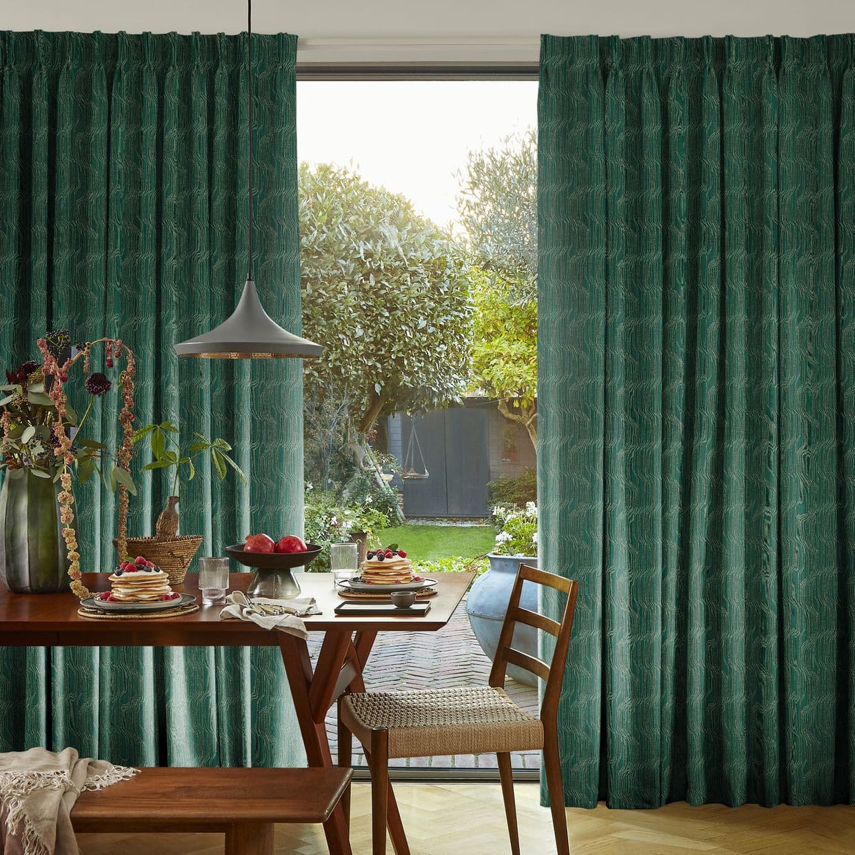 Floor-length emerald-green pleated curtains with textured pattern, parted centrally to frame the opening and admit soft daylight, revealing a dining nook with wooden table, pendant lamp, pancakes, and a sunlit garden view.