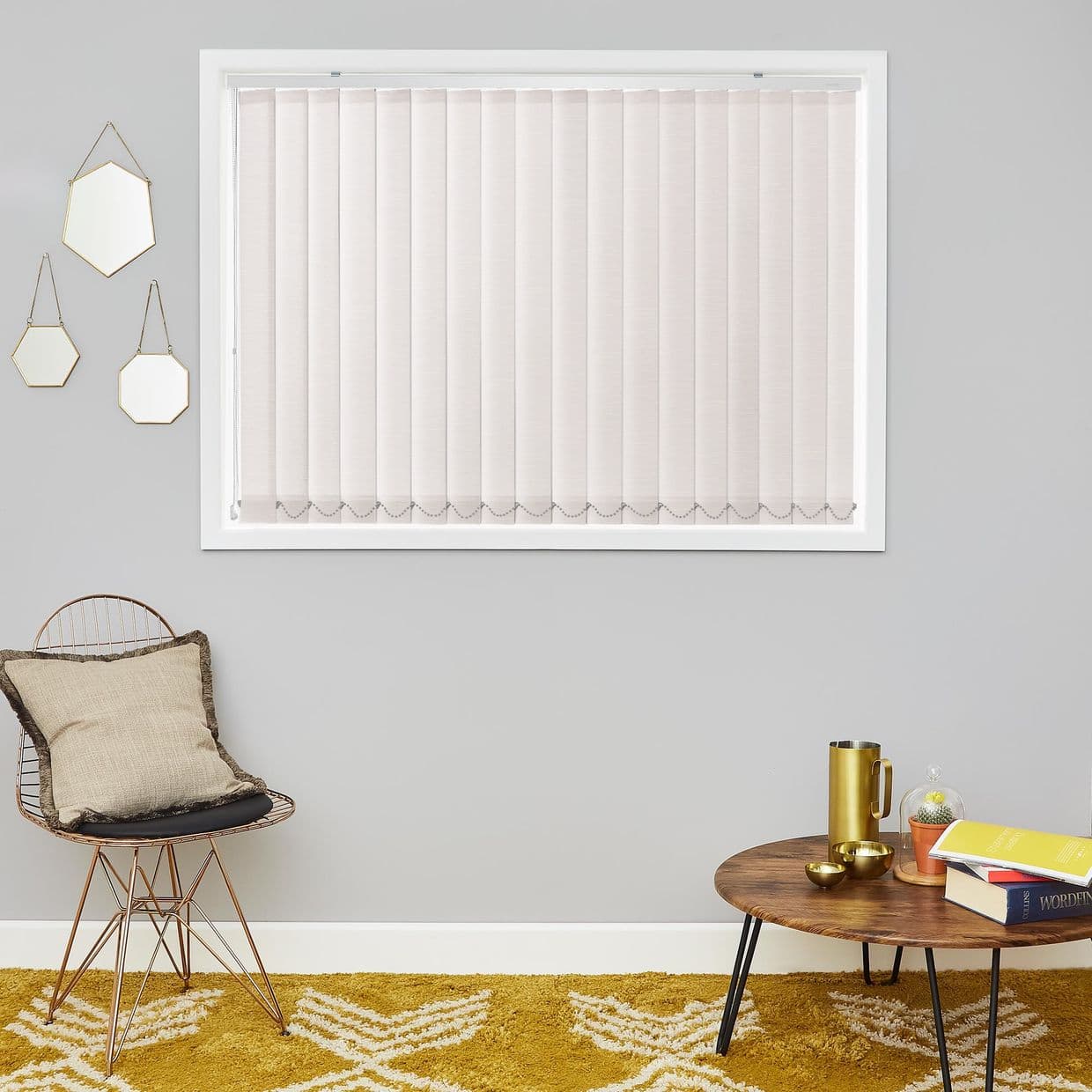 Beige vertical blinds covering a rectangular window, evenly spaced fabric slats with bottom beaded chain; closed, softly diffusing daylight; context: gray-walled living area with chair, wooden coffee table, mustard rug.