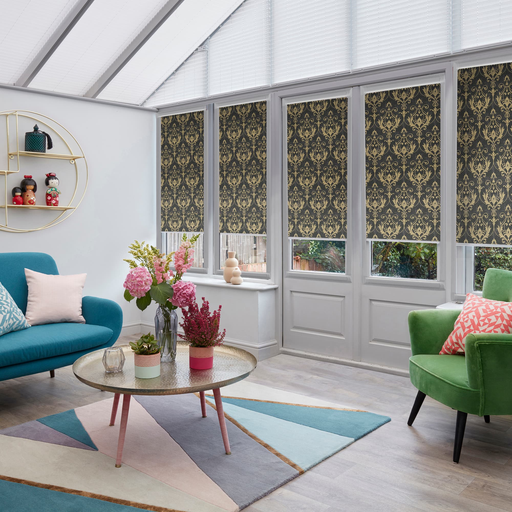Decorative dark roller blinds with gold damask pattern, lowered over tall conservatory windows, partially blocking light; set in a bright sunroom with glass roof, colorful sofas, coffee table and flowers.
