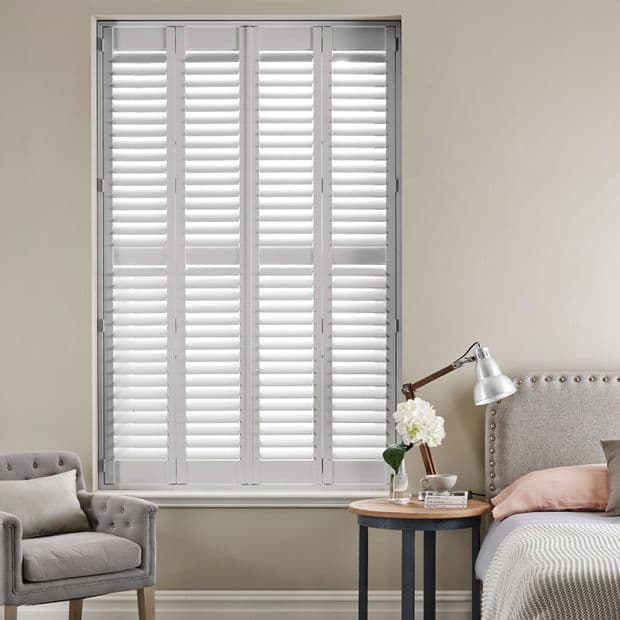 White louvered plantation shutters spanning a tall window, closed with slats slightly diffusing light; bedroom setting with upholstered headboard, bedside table with lamp and flowers, and an armchair.