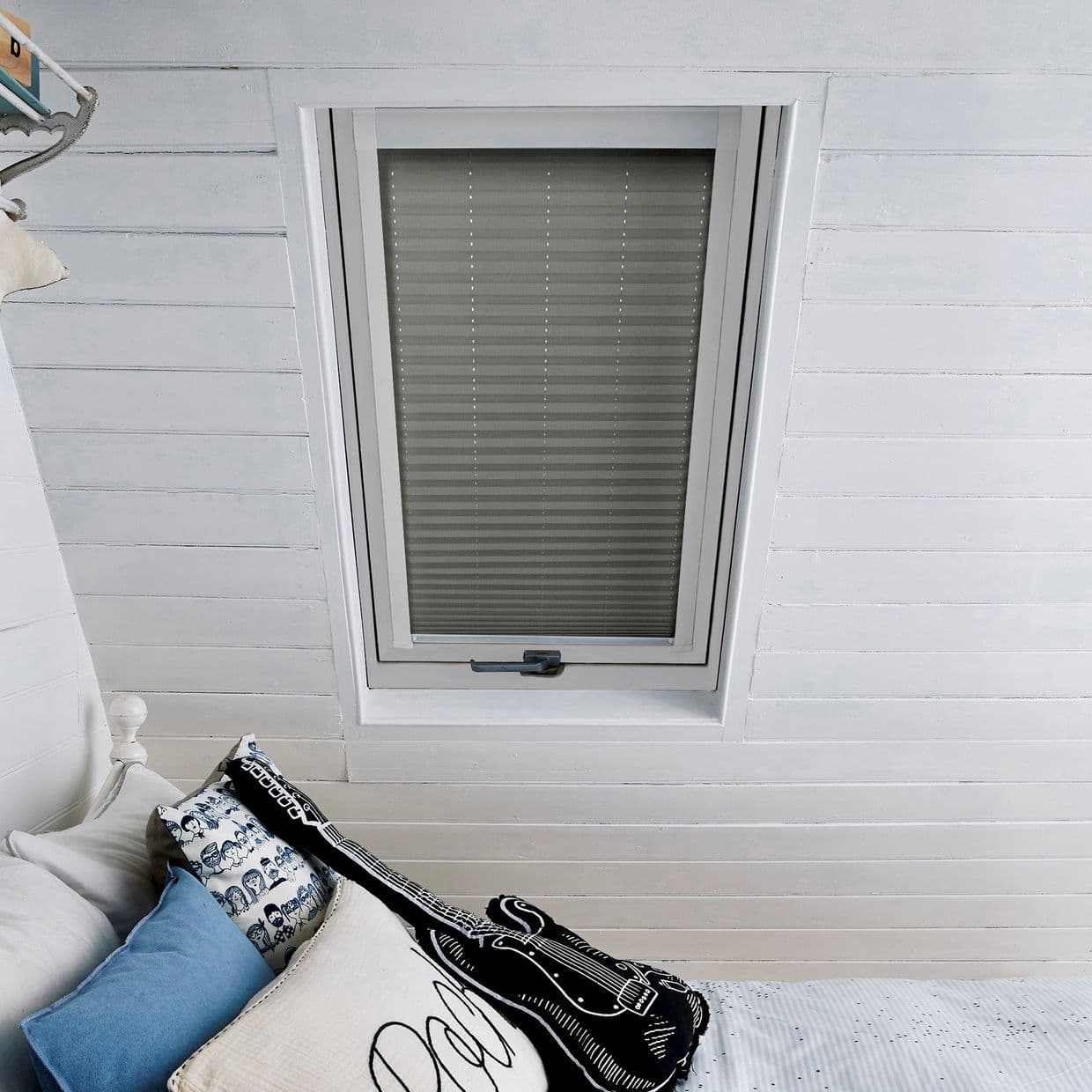 Pleated gray interior blind fully lowered over a rectangular skylight, blocking light; surrounded by white painted tongueâ€‘andâ€‘groove ceiling in a cozy attic bedroom with pillows and a guitar cushion.