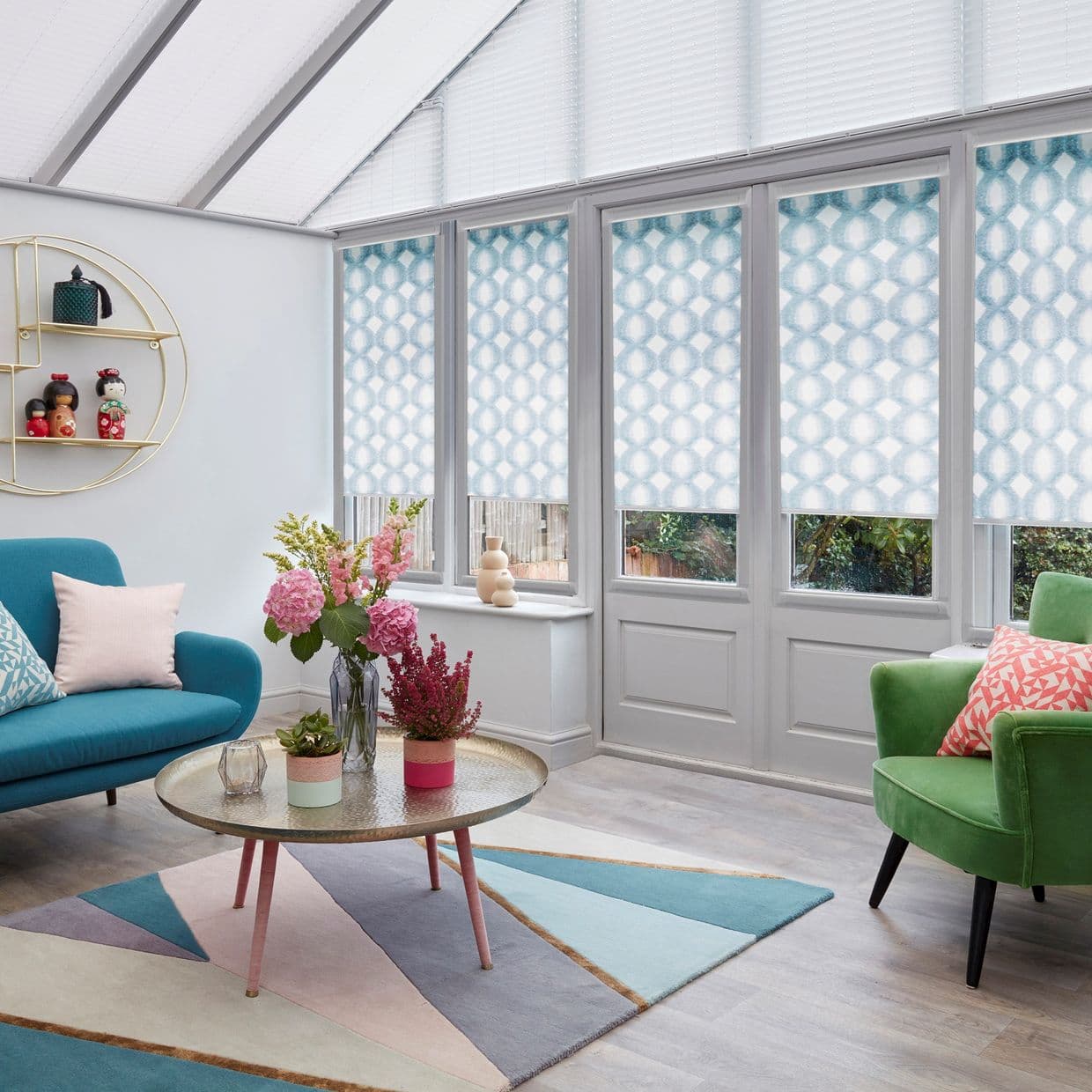 Blue-and-white geometric roller blinds on tall conservatory windows and doors, partially lowered and diffusing daylight; sunlit living space with pastel rug, teal sofa, green armchair, round coffee table and flowers.