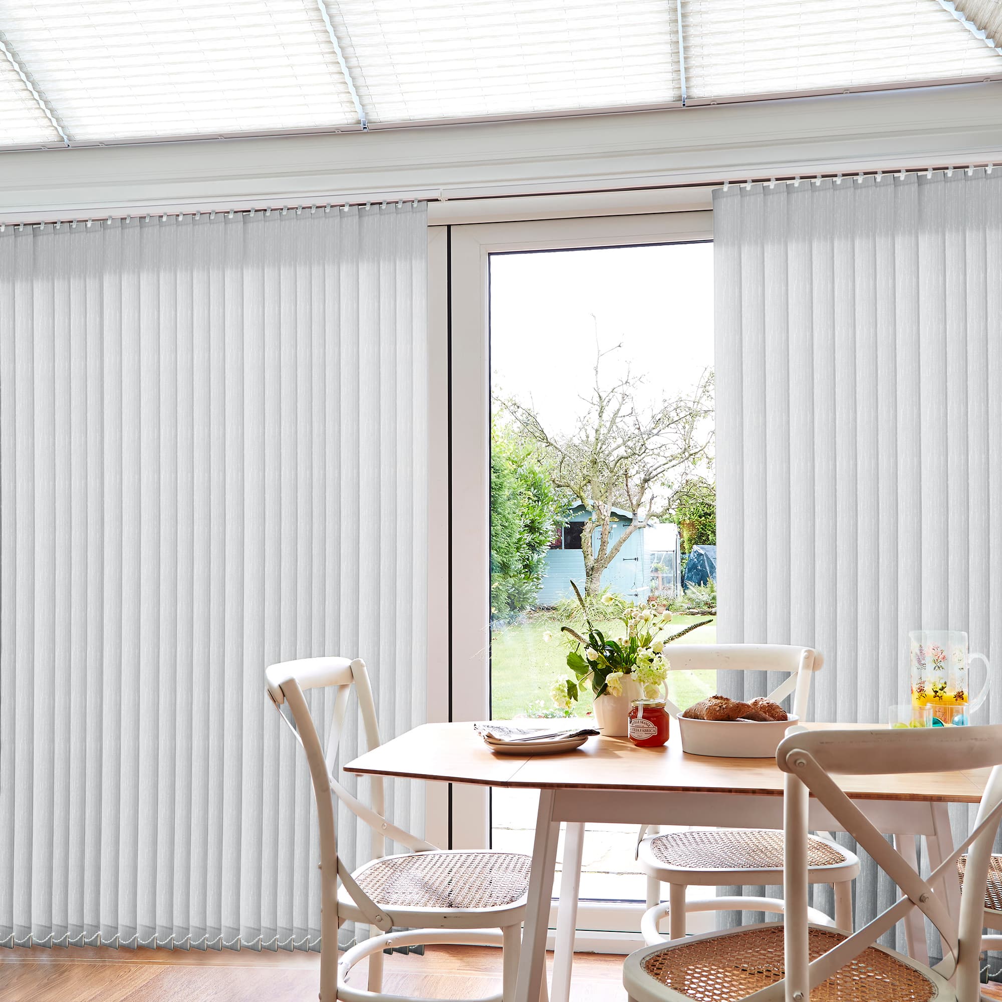White vertical blinds cover a glass patio door, mostly closed but parted centrally to filter daylight; view shows a sunlit dining table and chairs with a garden and shed beyond.