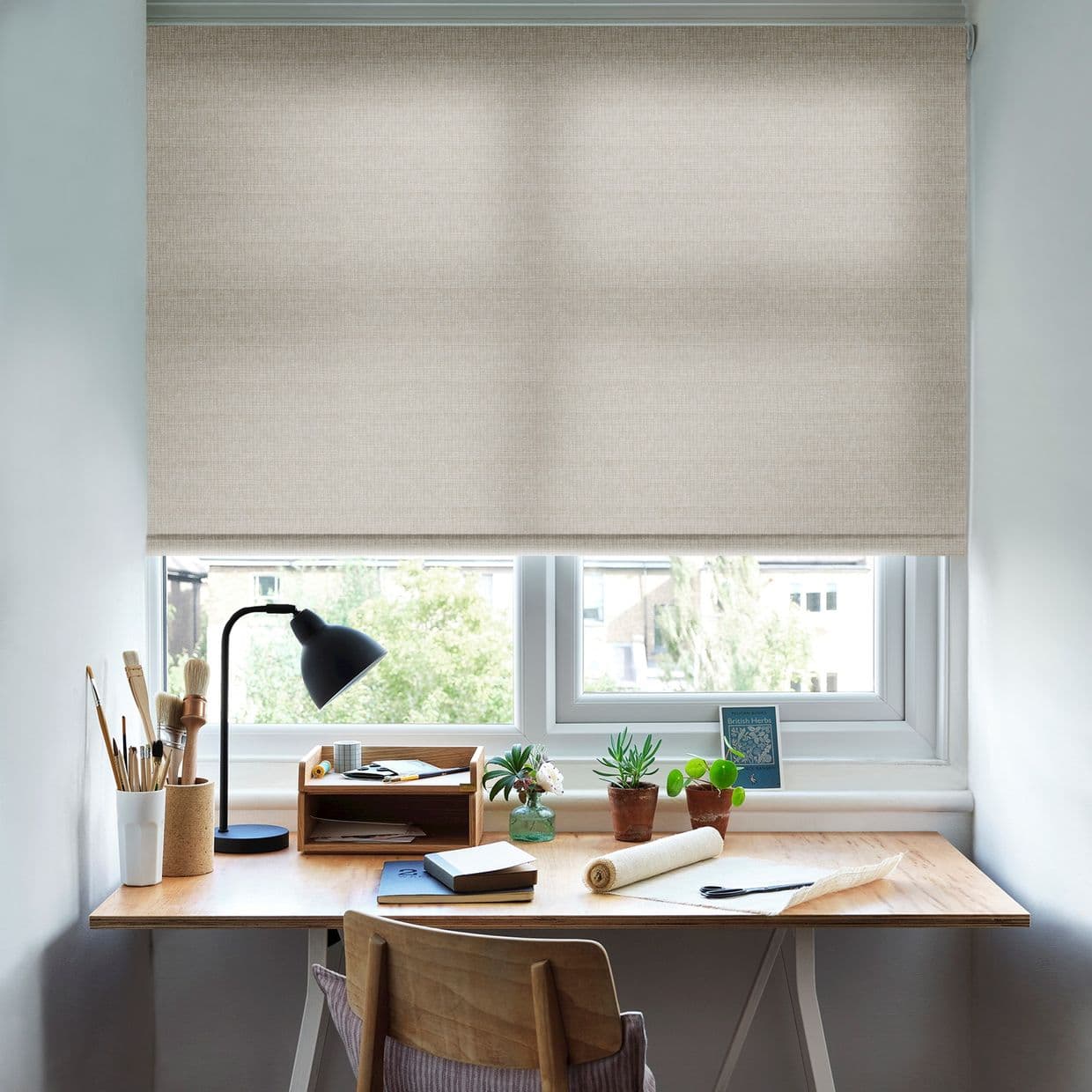 Beige fabric roller blind, fully lowered across two-pane window, softly diffusing daylight; above a wooden desk with lamp, brushes, notebooks and potted plants in a bright home workspace.