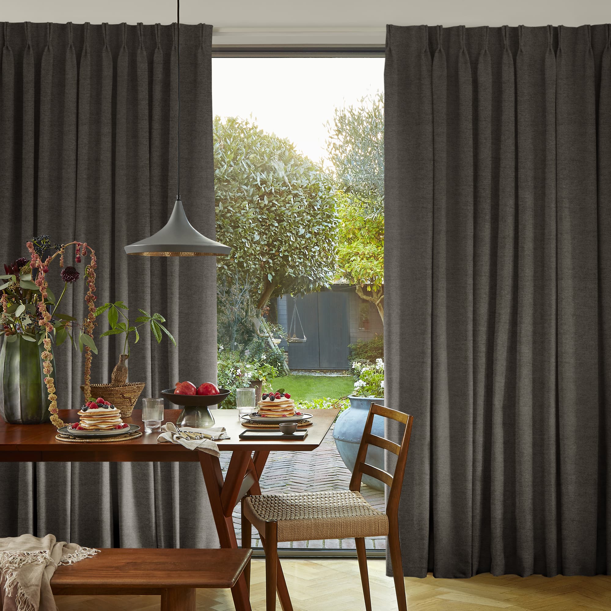 Floor-length pleated dark-gray curtains, partly drawn to frame a central opening, letting daylight into a dining room with wooden table set for breakfast, a chair, and a sunlit garden beyond.