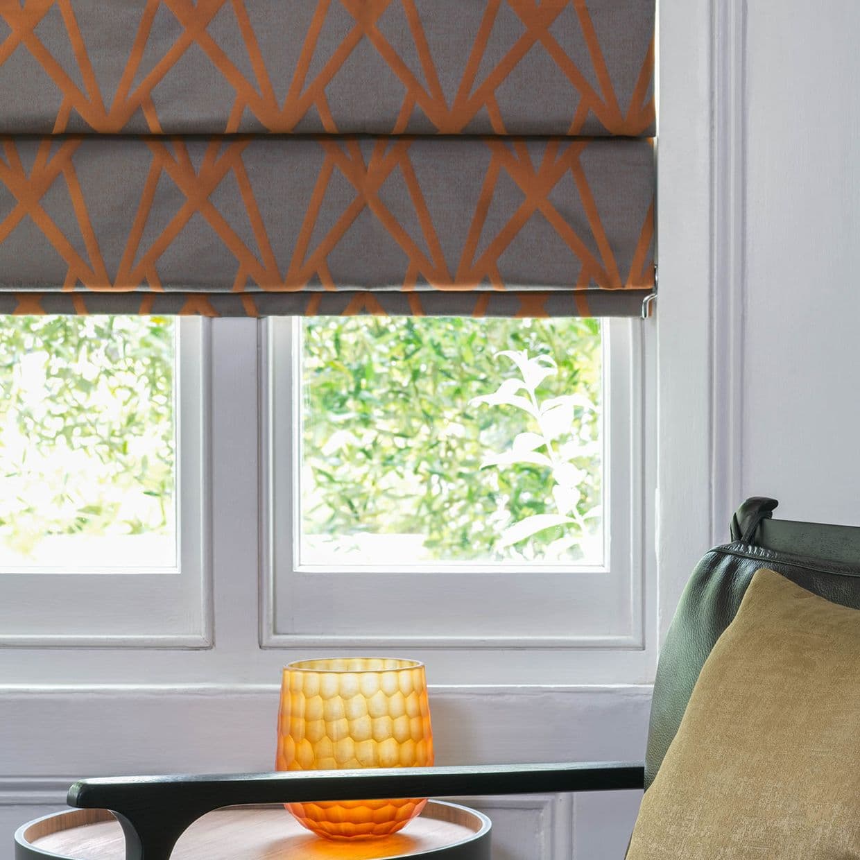 Roman blind, gray with orange geometric pattern, partially lowered, filtering daylight; over white-framed window with leafy green view, amber textured glass candle on a side table beside an upholstered armchair.
