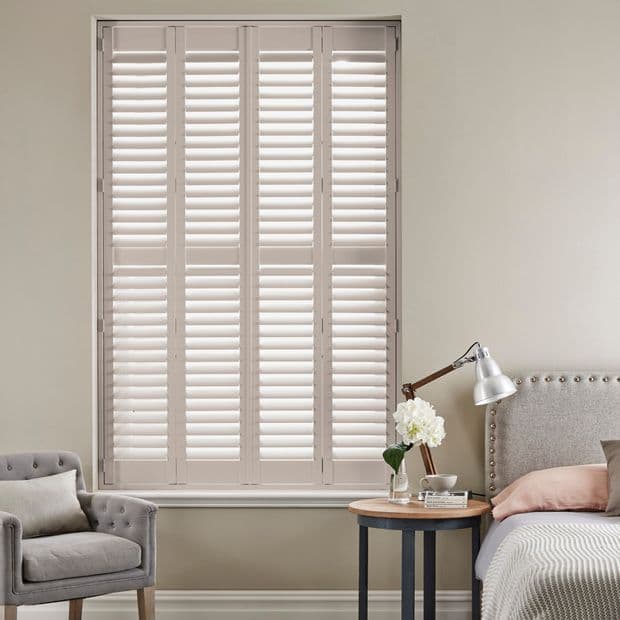 Cream-colored plantation shutters fill a tall window, fully closed with slats angled to filter soft daylight; set in a neutral bedroom with upholstered bed, round side table, lamp and armchair.