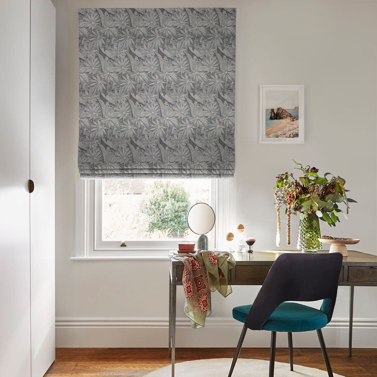 Grey patterned Roman blind, lowered with repeating leaf motif, partially filtering daylight through sash window; in bright neutral study with wooden desk, teal chair, round mirror and vase of flowers.