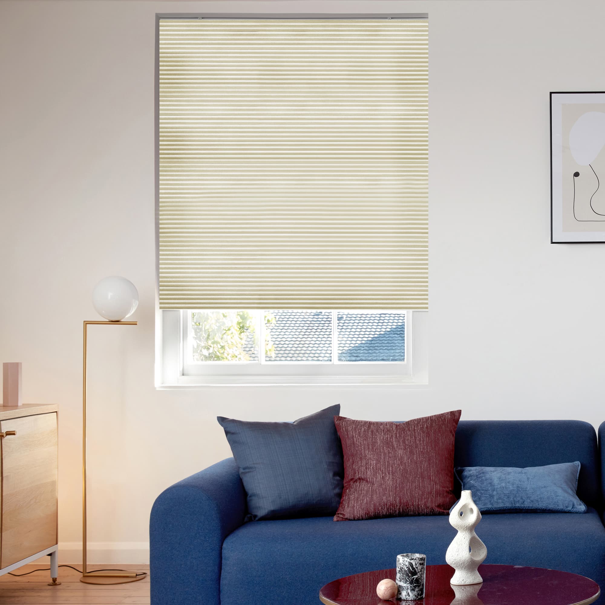 Beige pleated window blind, lowered with horizontal folds, filtering soft daylight and leaving a small bottom gap; modern living room with blue sofa, cushions, floor lamp and abstract wall art.