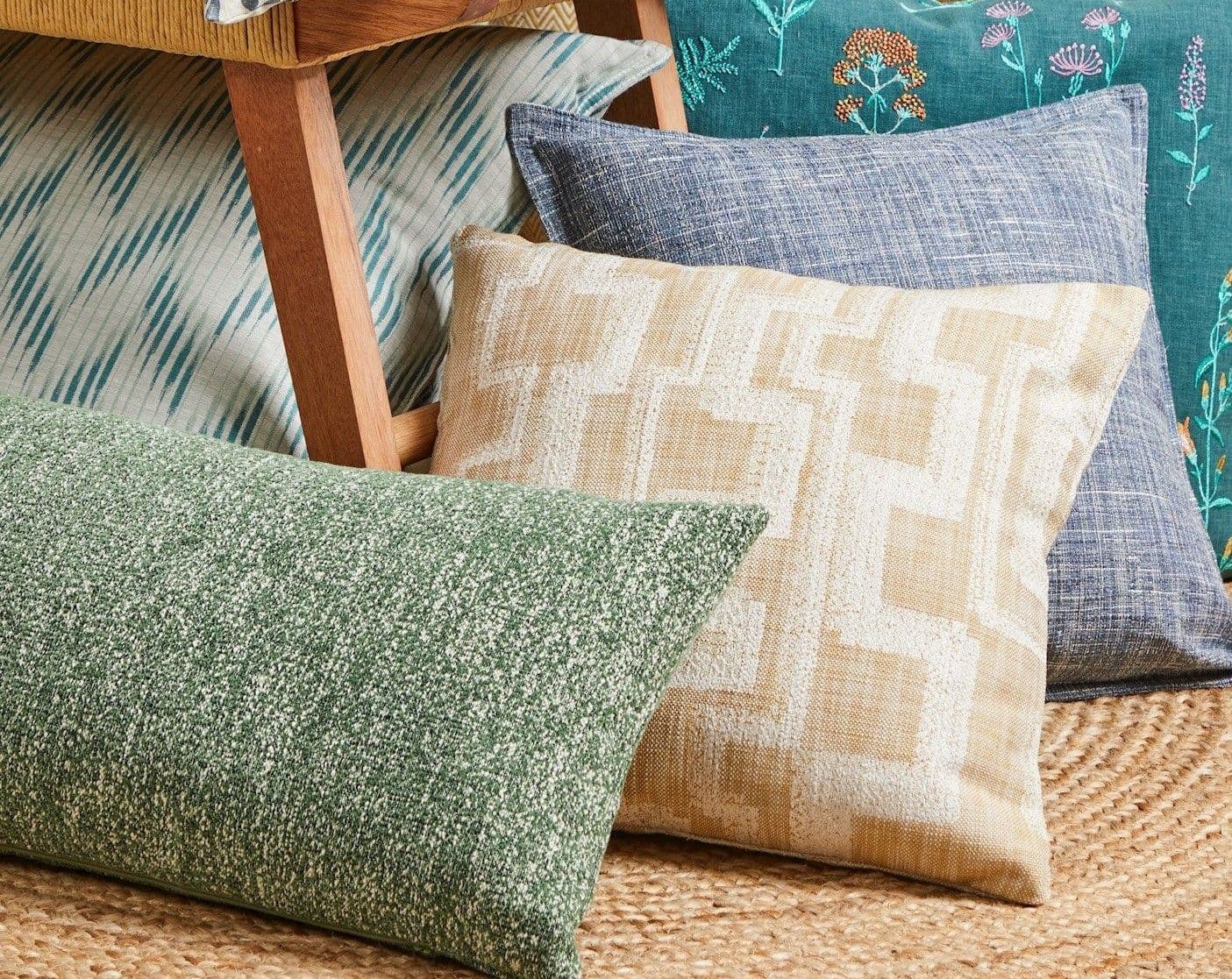 Decorative cushions (green lumbar, beige geometric, blue textured, floral teal) arranged leaning against a wooden chair leg, resting on a woven jute rug in a softly lit cozy seating corner.