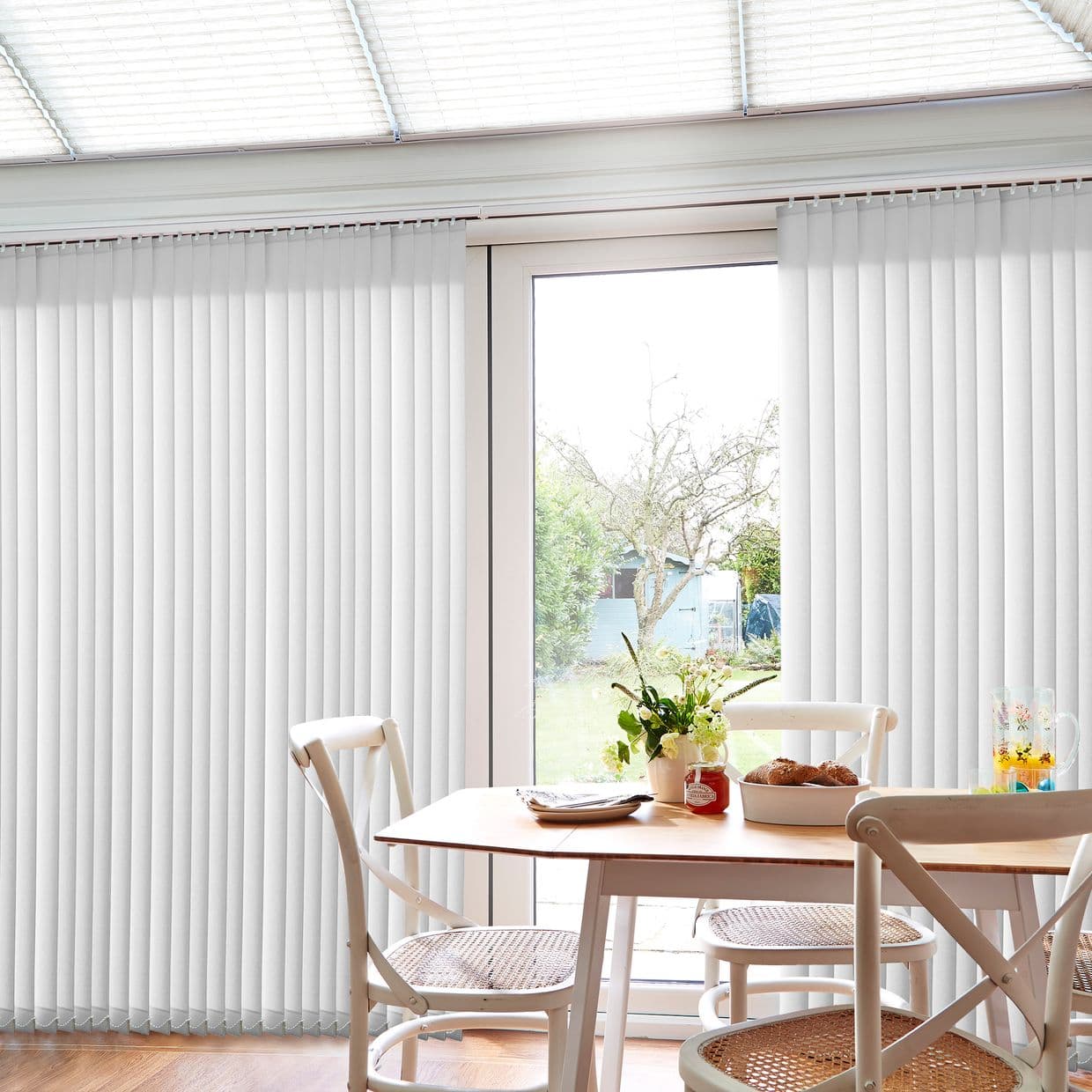 White vertical blinds (floor-to-ceiling slats on a ceiling track), mostly closed but parted at center to reveal a glass patio door; daylight filters into a small dining area and garden.