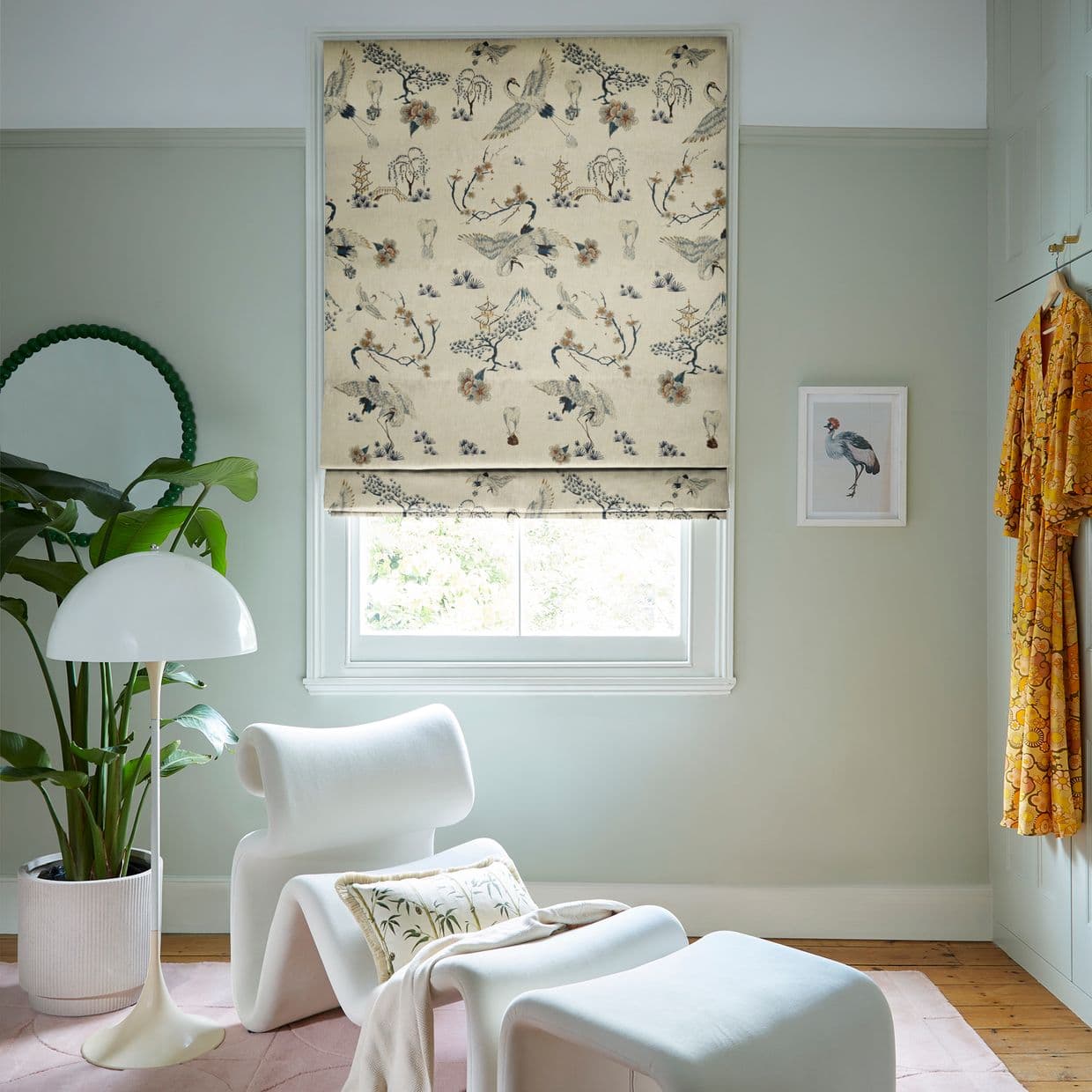 Printed Roman blind featuring cranes and blossoms, lowered over the window, filtering daylight into a pale-green reading nook with white lounge chair, potted plant, floor lamp and framed bird artwork.