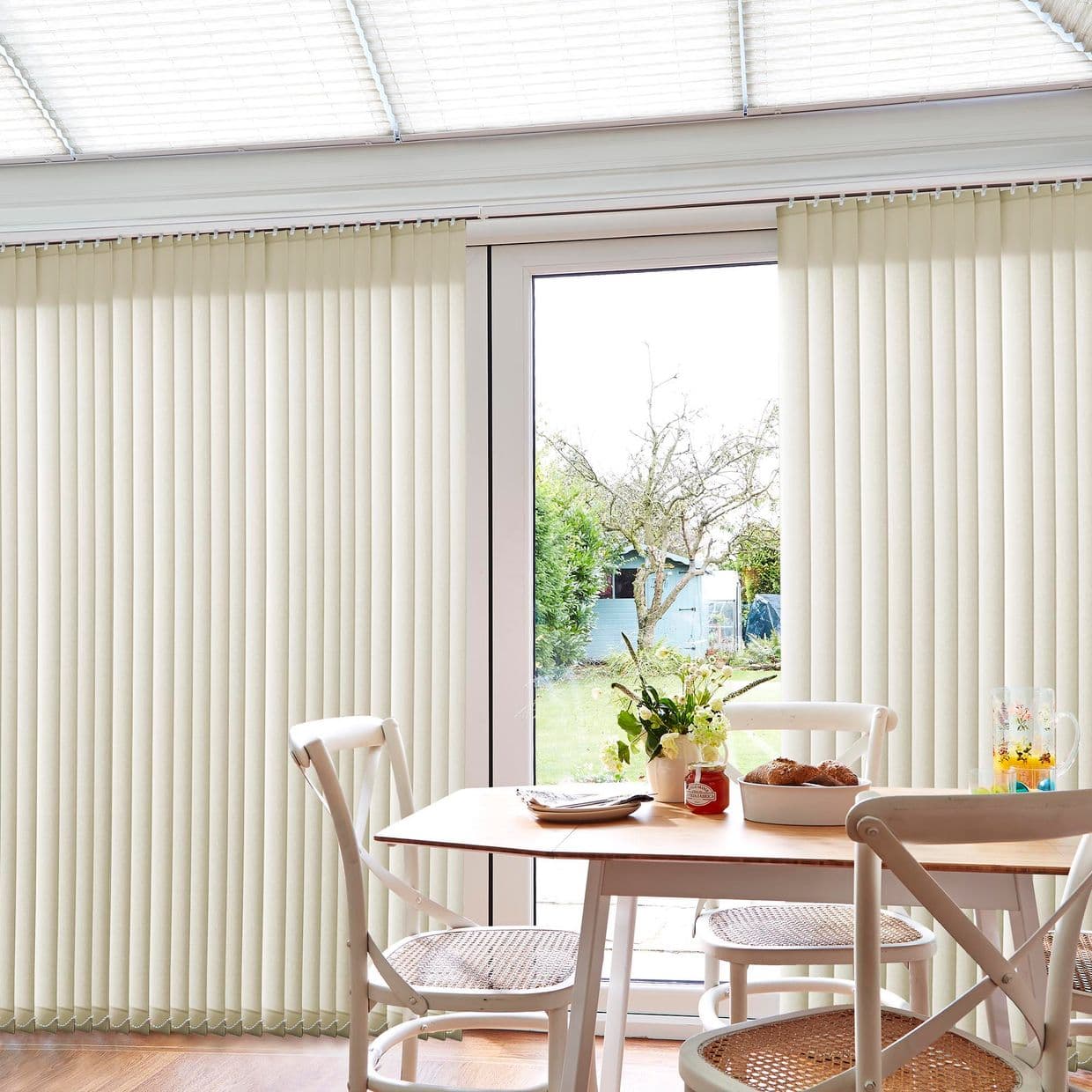 Cream vertical blinds cover a sliding glass door, mostly closed with a narrow central gap letting daylight through; a dining nook with wooden table, chairs, flowers, pastries and backyard view.