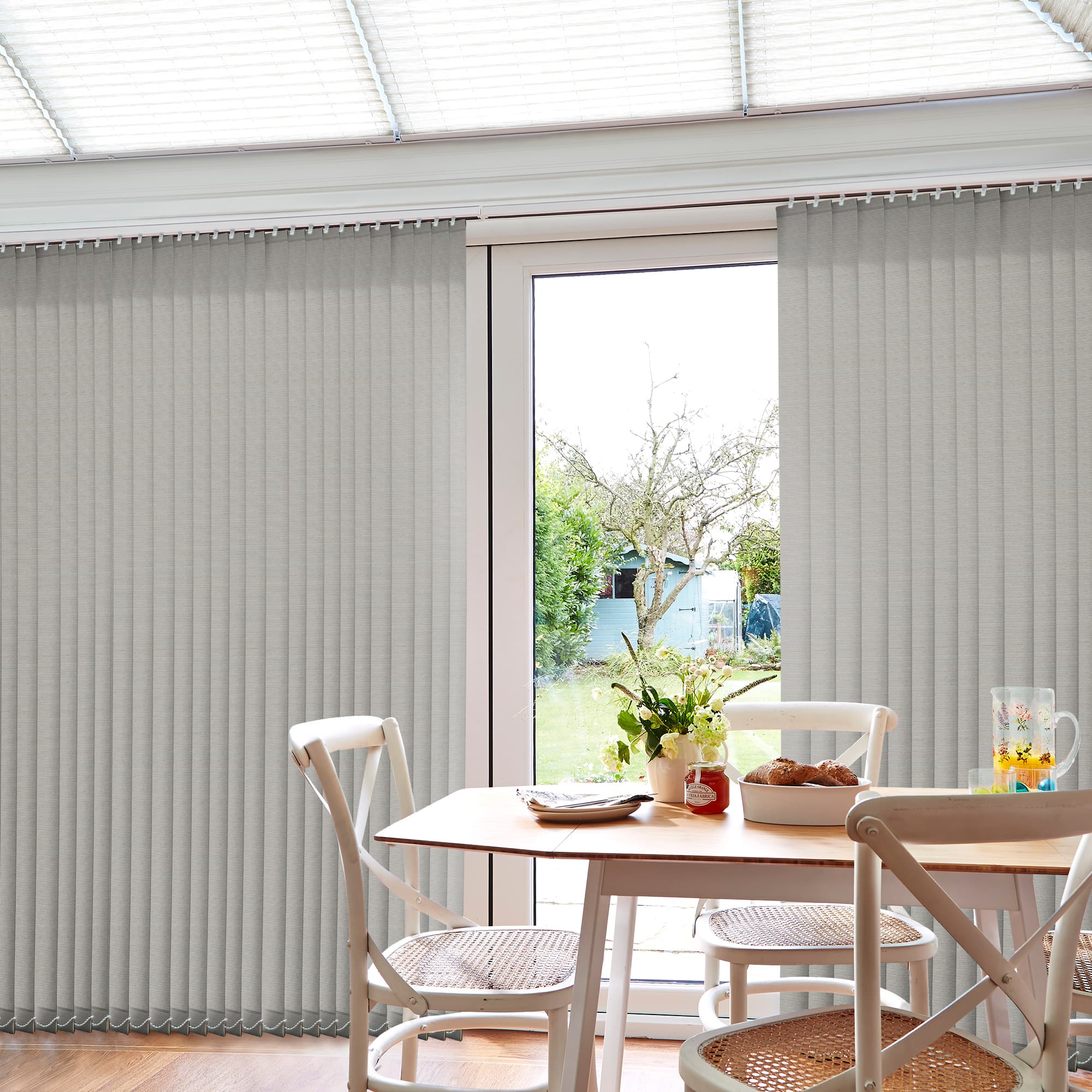 Grey fabric vertical blinds, floor-to-ceiling with slim slats and visible headrail; mostly closed but parted centrally, filtering daylight into a bright dining nook with wooden table, chairs, and a garden view.