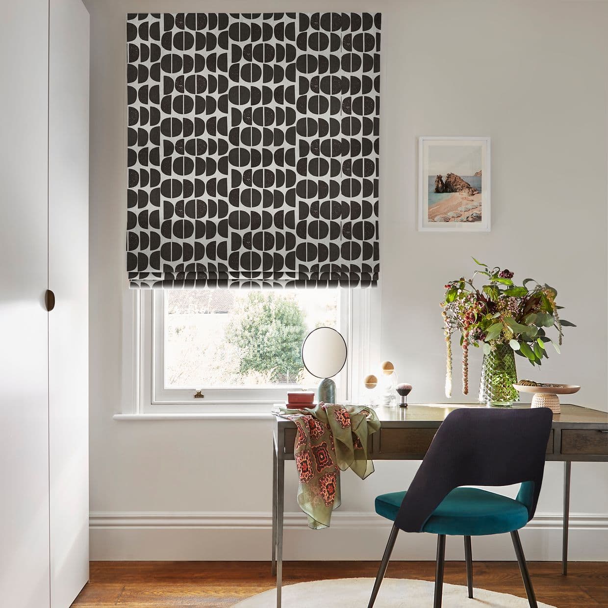 Black-and-white geometric Roman blind, lowered mid-window with repeating semi-circle motif; partially closed, filtering daylight over a desk with chair, mirror and vase of flowers in a pale home office.