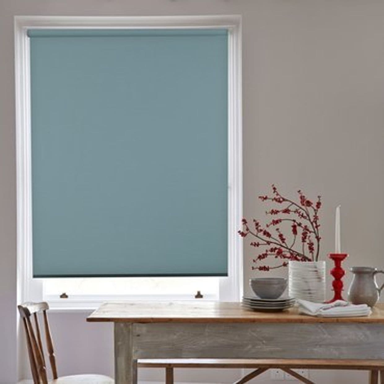 Teal roller blind, fully lowered with smooth fabric, blocking light; over a white-framed window above a rustic wooden table with stacked dishes, vase of red branches and a red candlestick.