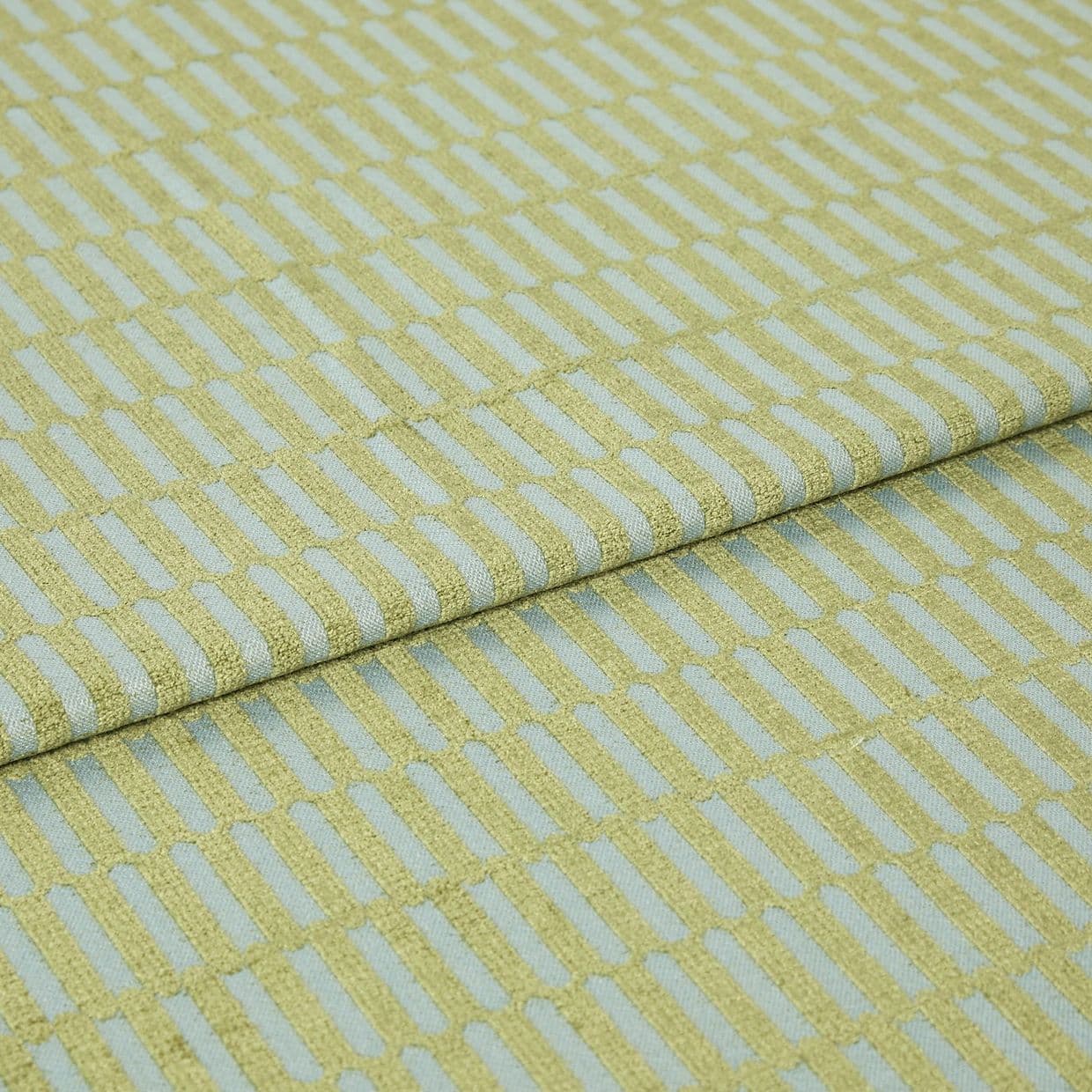 Textile fabric with raised yellowâ€‘green rectangular grid pattern, folded once along a horizontal seam and overlapping, close-up studio-lit sample on a pale blue ground, showing textured weave.