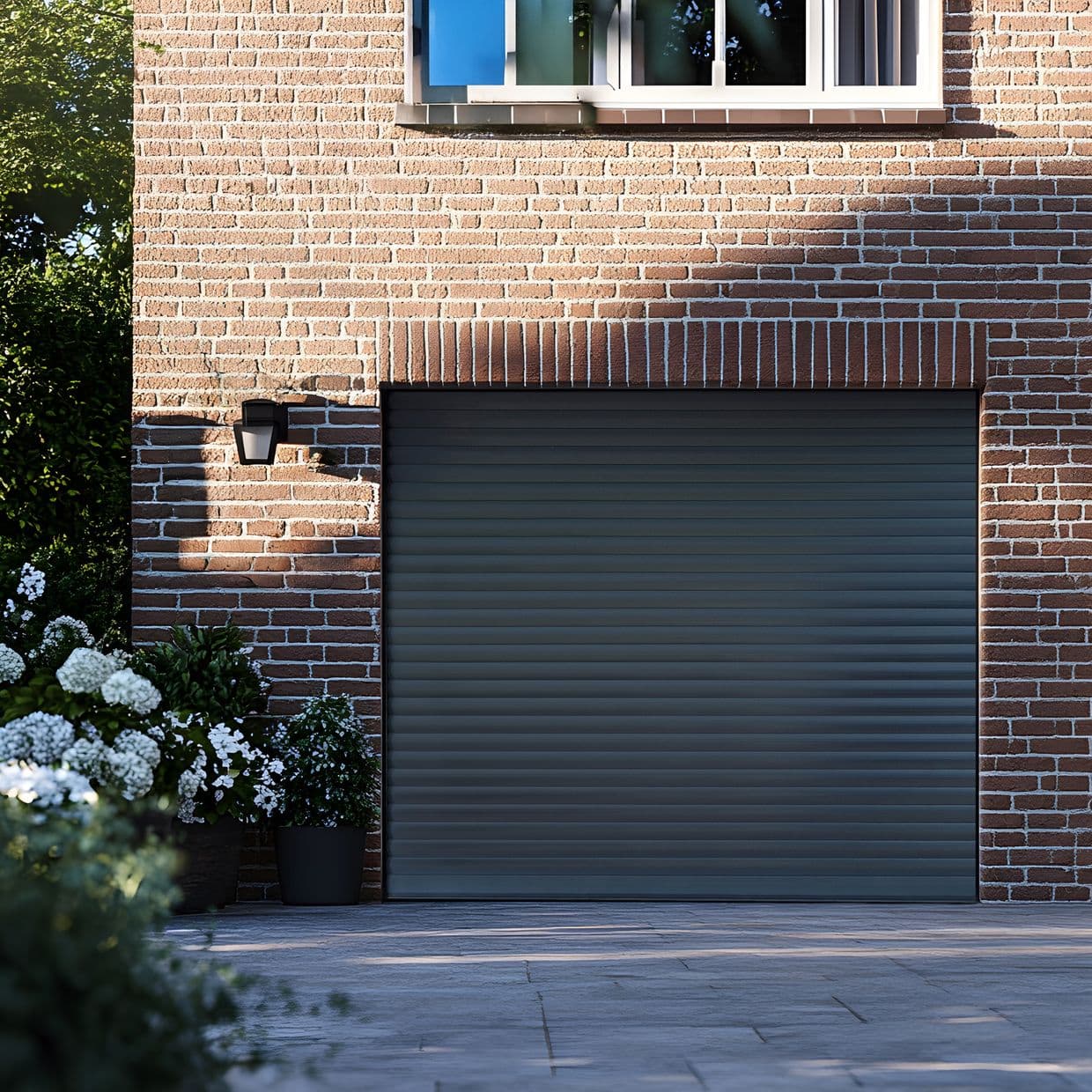 Horizontal slatted metal roller shutter covering garage opening, closed and blocking view; set in red brick wall beneath a window, with exterior lamp, potted white flowers and soft afternoon light.