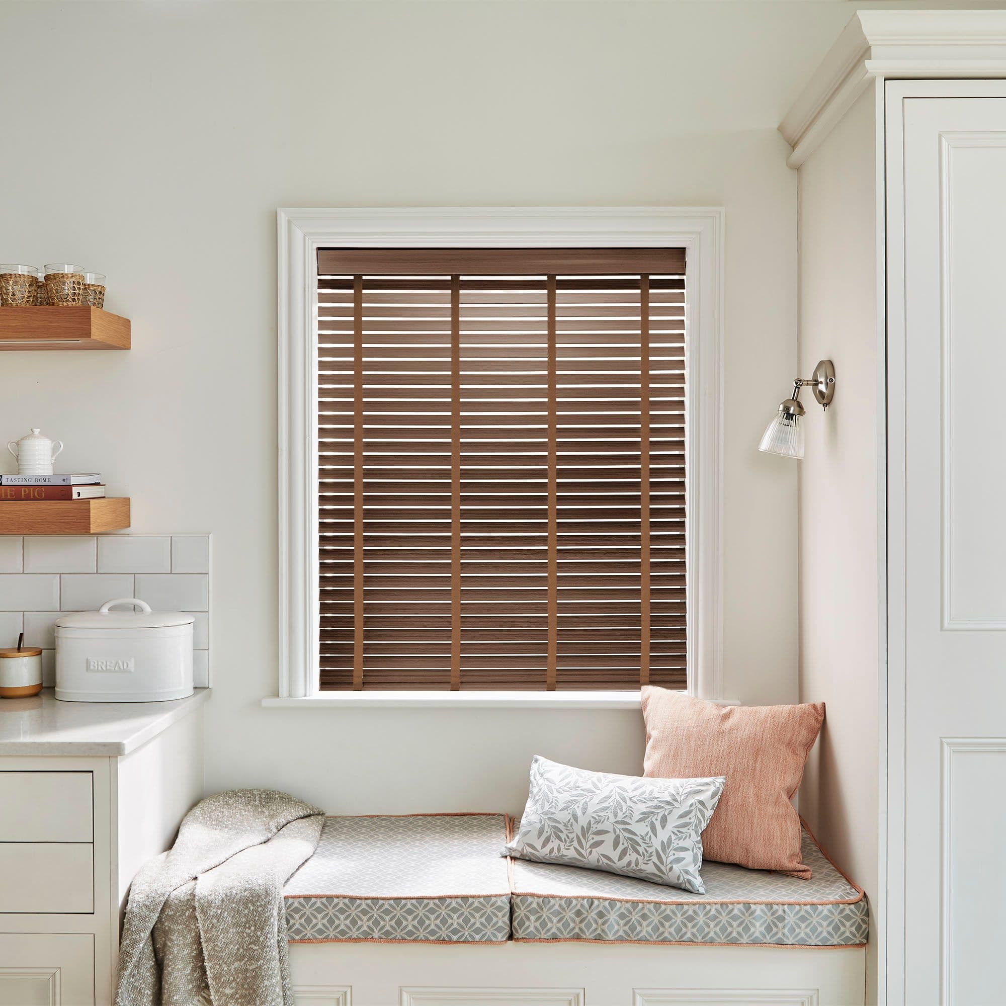 Wooden horizontal slat blinds lowered over the window, closed and gently filtering daylight; set in a neutral-toned kitchen alcove with a built-in cushioned bench, pillows, shelves and a wall sconce.