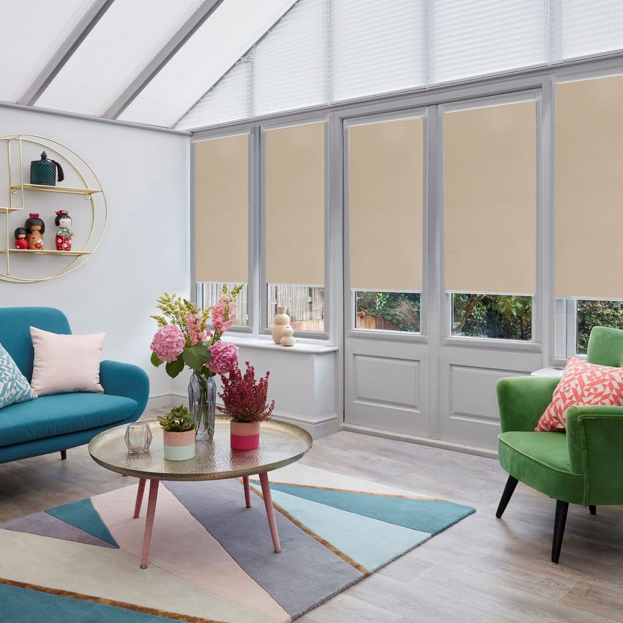Beige roller blinds, lowered halfway across tall conservatory windows and doors, filtering daylight; bright sunroom living area with teal sofa, green armchair, round coffee table with flowers and garden glimpses.