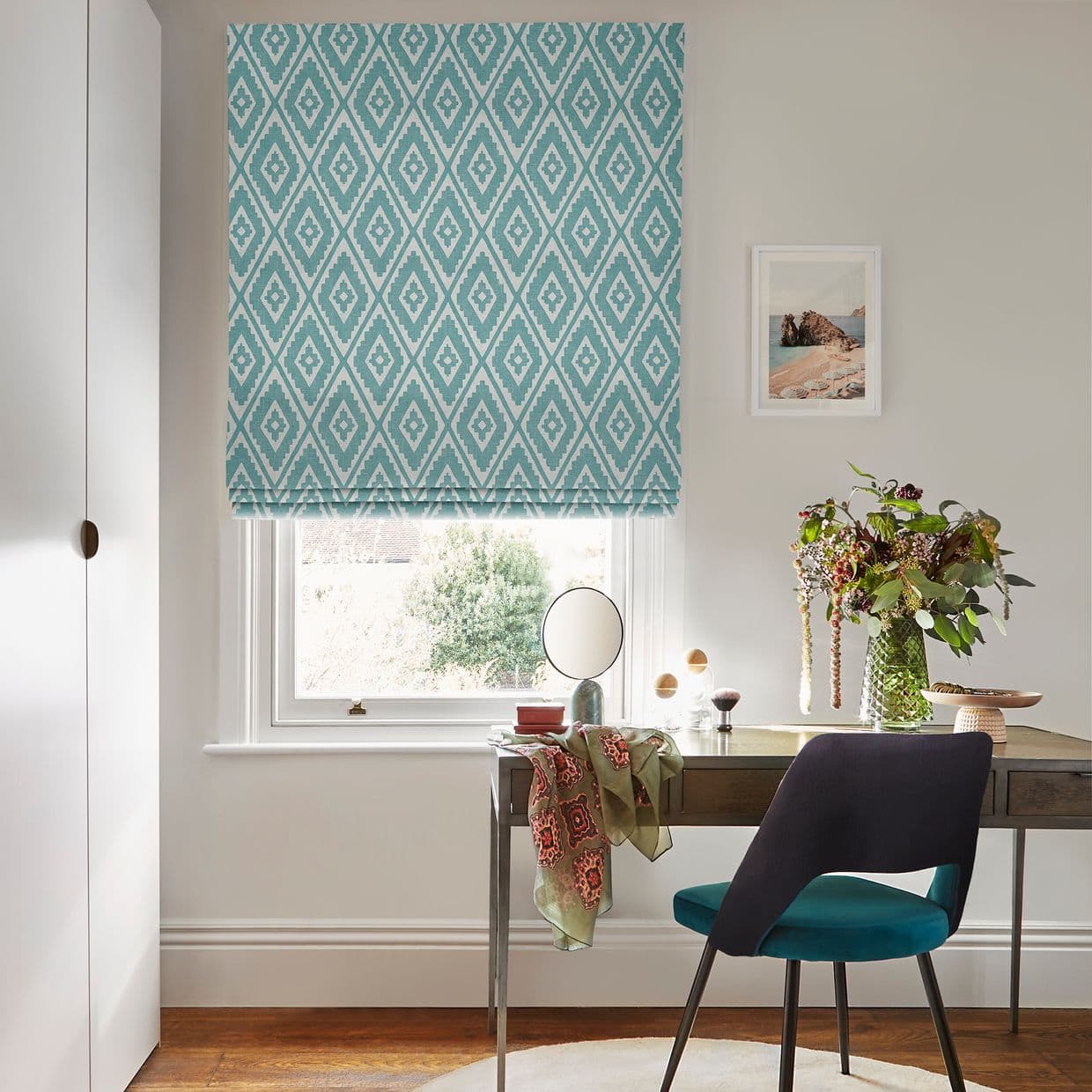 Teal-and-white diamond-patterned Roman blind lowered over a window, filtering daylight; in a small home office, a desk with mirror, vase of flowers, scarf-draped surface and teal-upholstered chair.