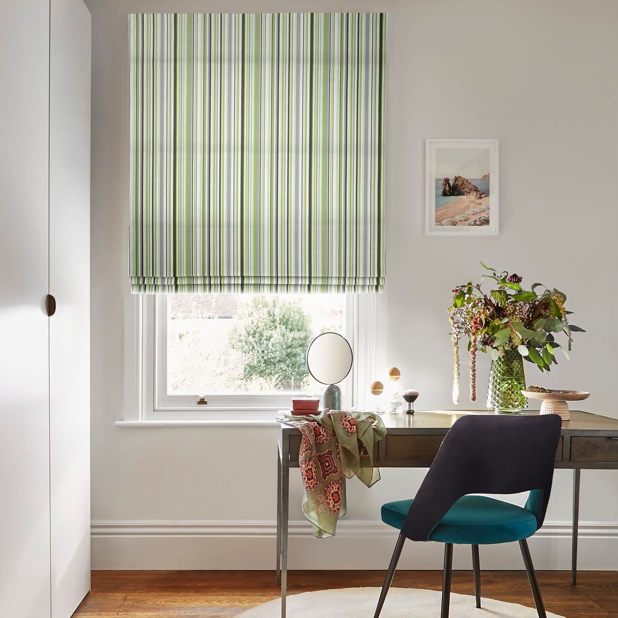 Striped Roman blind (green, grey, white vertical stripes) lowered, filtering soft daylight through a sash window; home desk with teal chair, vase of flowers, scarf-draped tabletop and framed print.