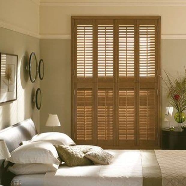 Wooden full-height plantation shutters covering a tall windowâ€”four hinged panels with louvered slatsâ€”closed and filtering warm daylight into a neutrally toned bedroom with bed, lamps, and wall decor.