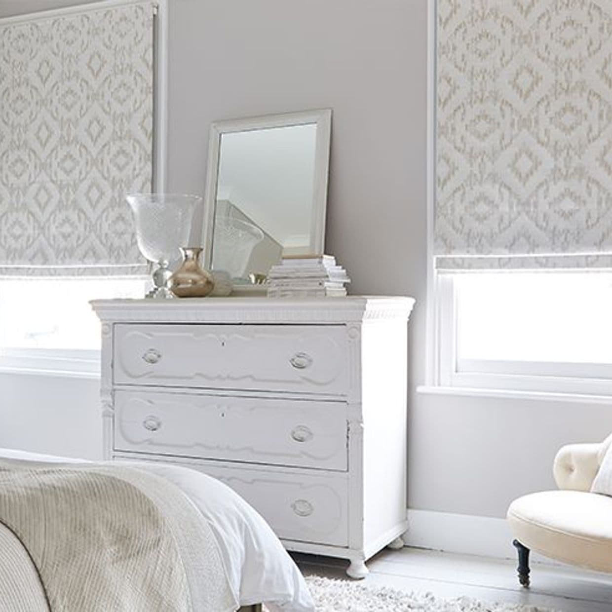 Patterned fabric Roman shades, lowered and filtering daylight through pale geometric motifs; positioned over two windows above a white distressed three-drawer dresser â€” neutral bedroom with mirror, vases, soft lighting.