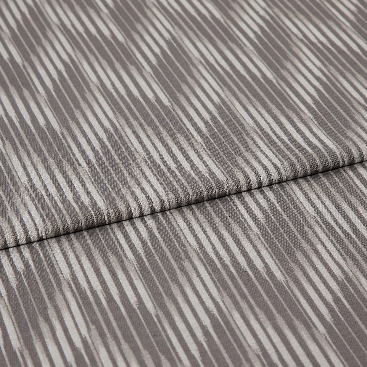 Folded curtain fabric sample with narrow, irregular vertical grey and off-white stripes; folded edge across center, shown close-up on a neutral surface under soft, diffuse lighting.
