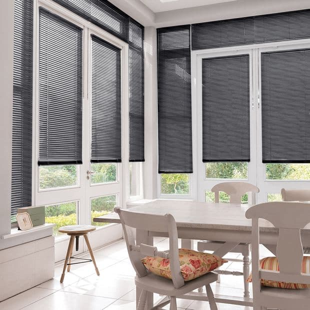 Dark horizontal slatted blinds covering tall bay windows, mostly lowered to mid-height. They filter and block light. Bright sunroom/dining area with white table and chairs, garden visible through lower panes.