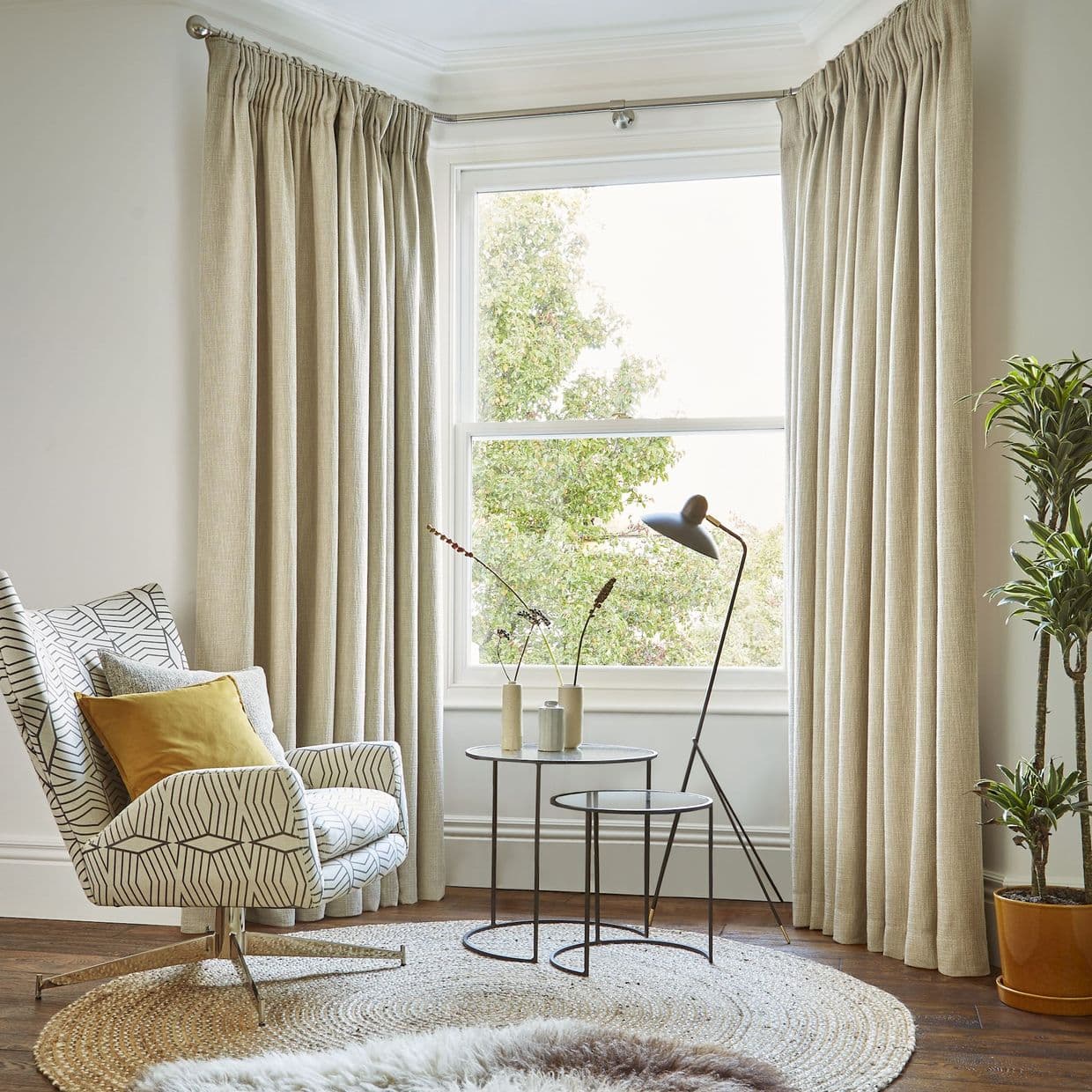 Beige pleated curtains on a curved rod, hung floor-to-ceiling at window edges; they frame and softly filter daylight through the window, in a living-room corner with patterned armchair, lamp, rug.