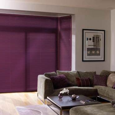 Purple horizontal slatted blinds cover a bay window, fully closed and filtering light into a living room with an L-shaped olive sofa, low coffee table, wooden floor, and framed art.