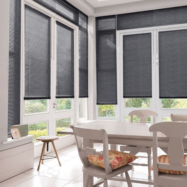 Dark gray horizontal-slat Venetian blinds cover tall corner windows and French doors, mostly lowered and filtering daylight with slight bottom openings; sunlit dining area with table, chairs and garden view.