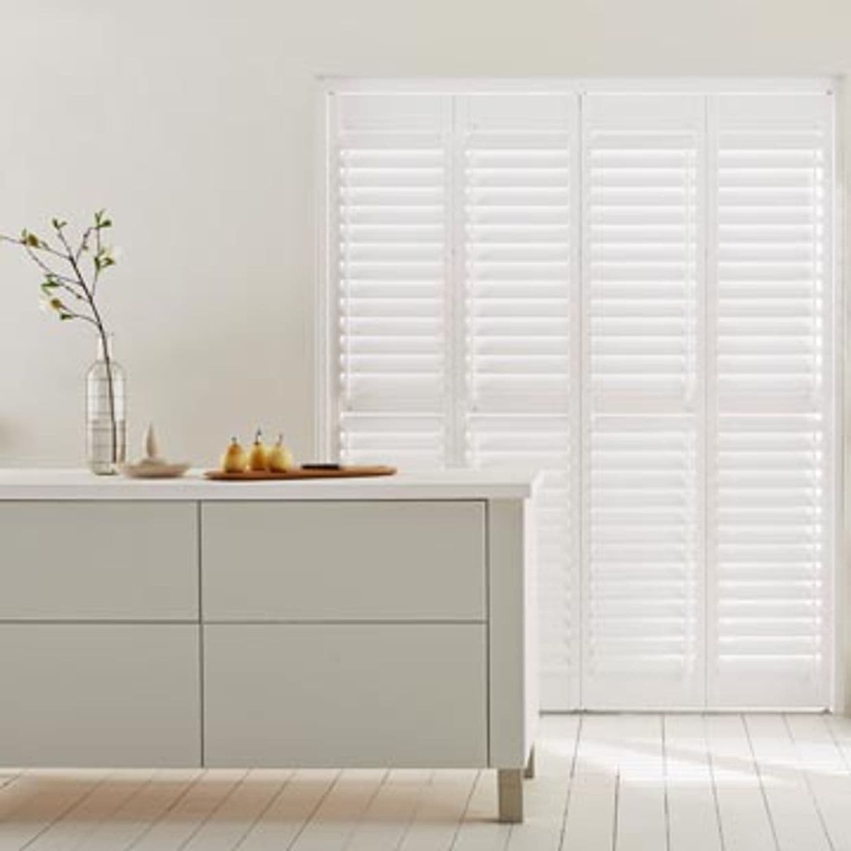 White plantation shutters across a tall window/door, fully closed with horizontal slats filtering bright daylight, in a minimalist kitchen with an island, vase with branch, wooden tray and light floor.