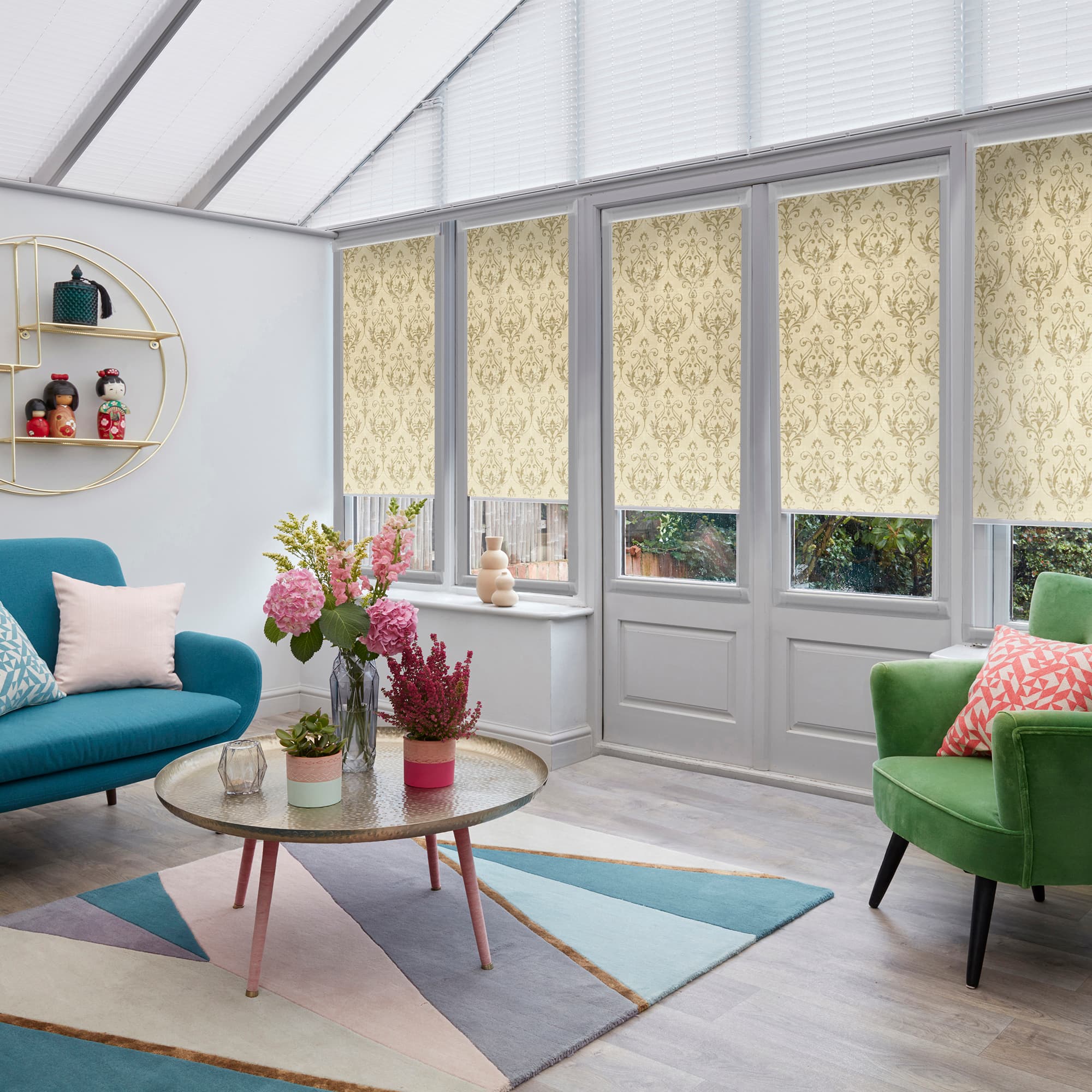 Patterned roller blinds (cream damask) cover tall rectangular windows and glass door, mostly lowered and filtering daylight; in a bright conservatory-style living room with teal sofa, green armchair, floral centerpiece.
