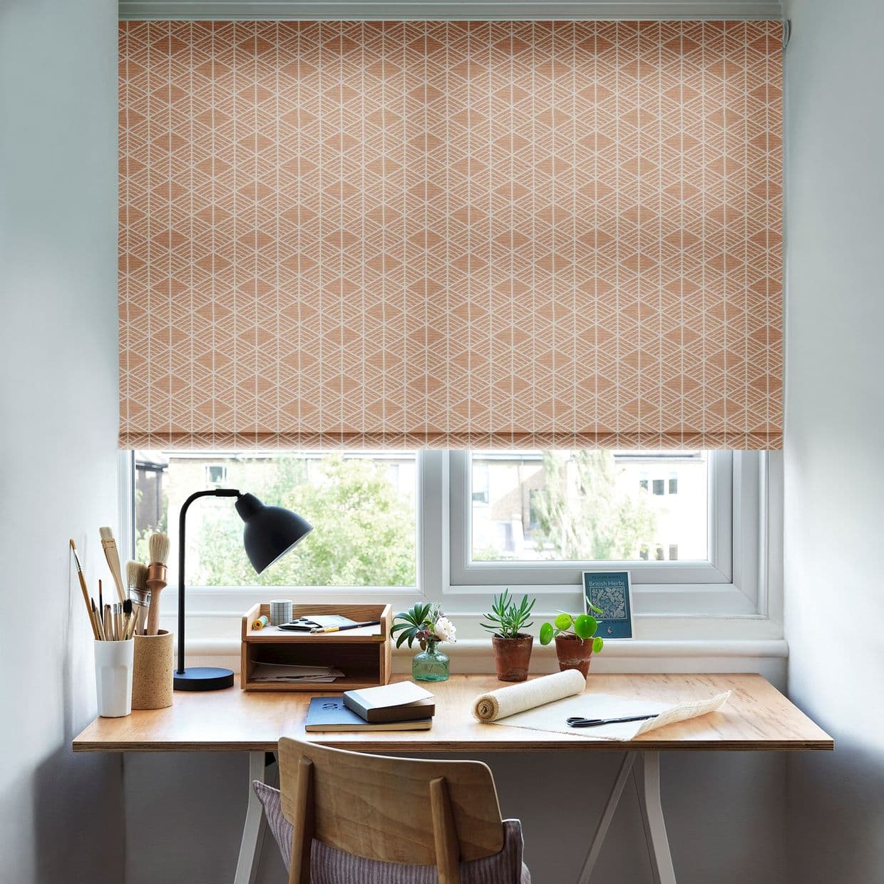 A peach roller blind with white geometric lattice, lowered to just above the sill, filtering daylight; above a tidy wooden desk with lamp, small plants and art supplies, suburban view.