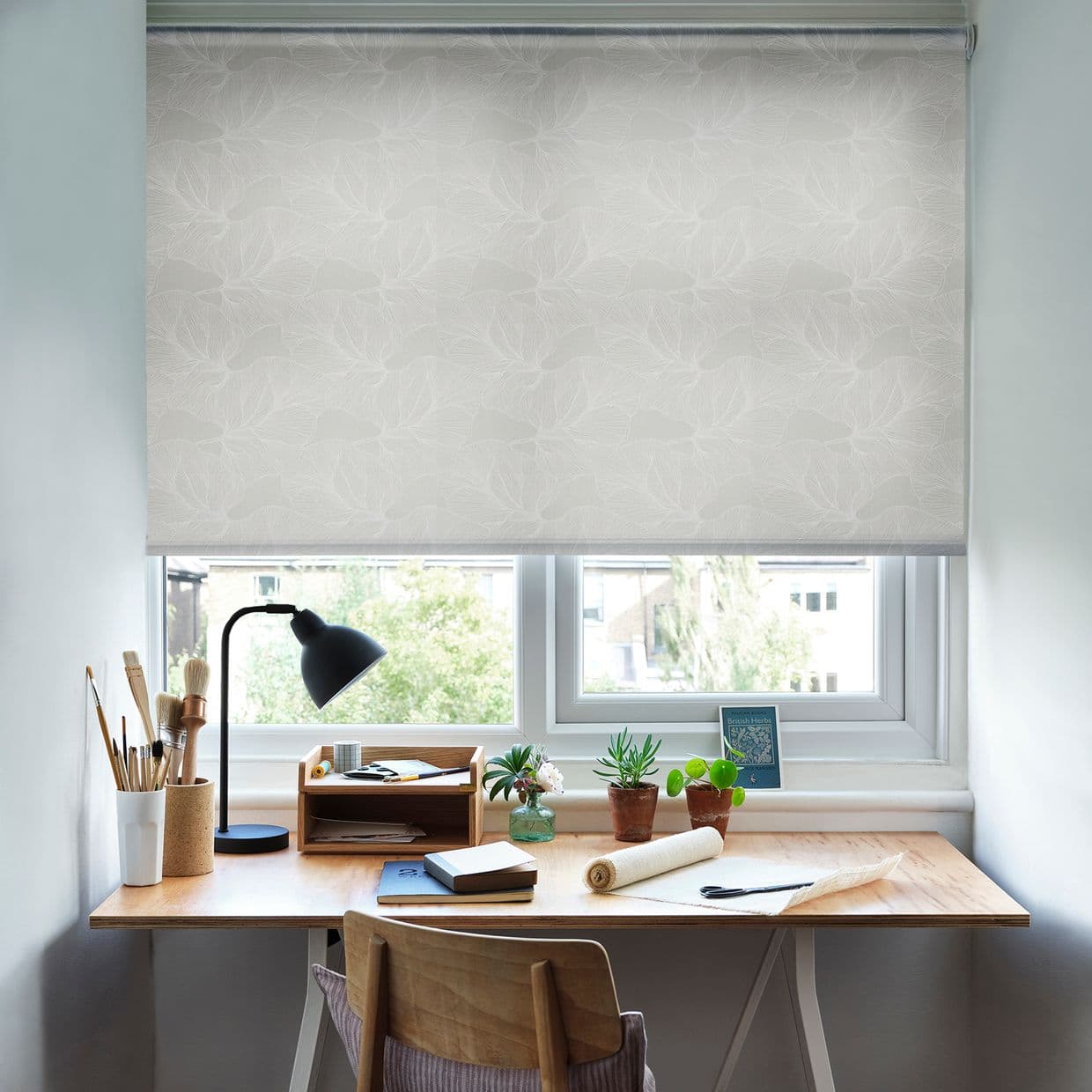 A pale floral roller blind pulled down across the window, softly filtering daylight; below, a wooden desk workspace with lamp, books, brushes and small potted plants, suburban view.