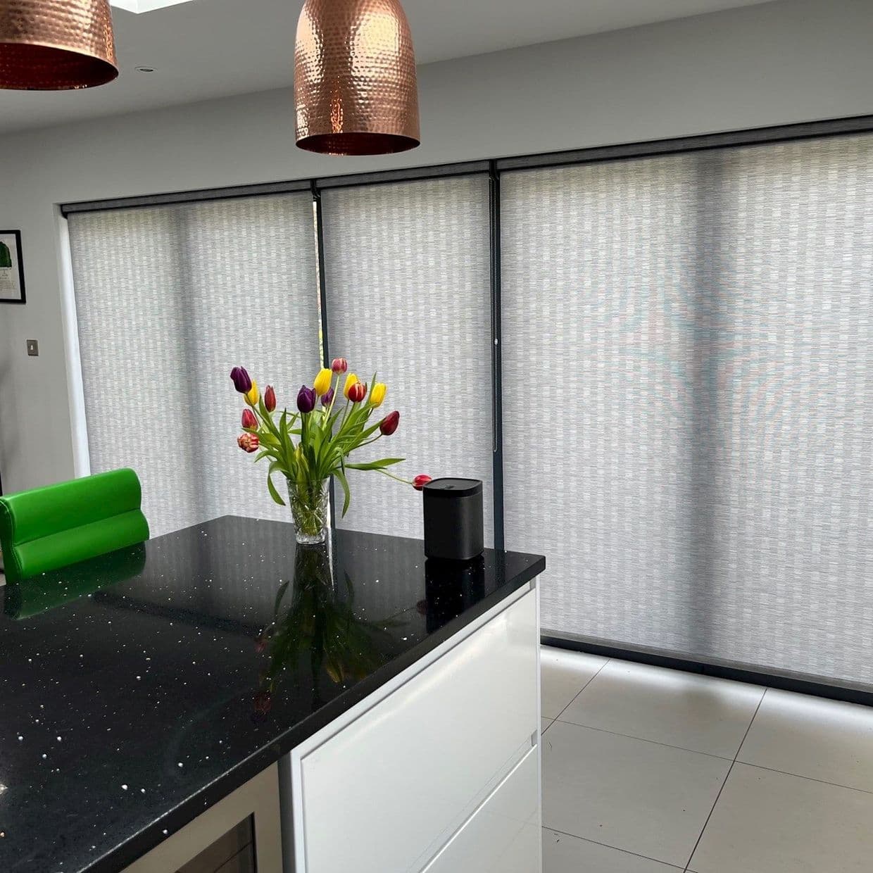 Light-gray patterned roller blinds span three sliding panels, fully lowered and diffusing daylight; modern kitchen with black island countertop, vase of tulips, copper pendant lights and tiled floor.