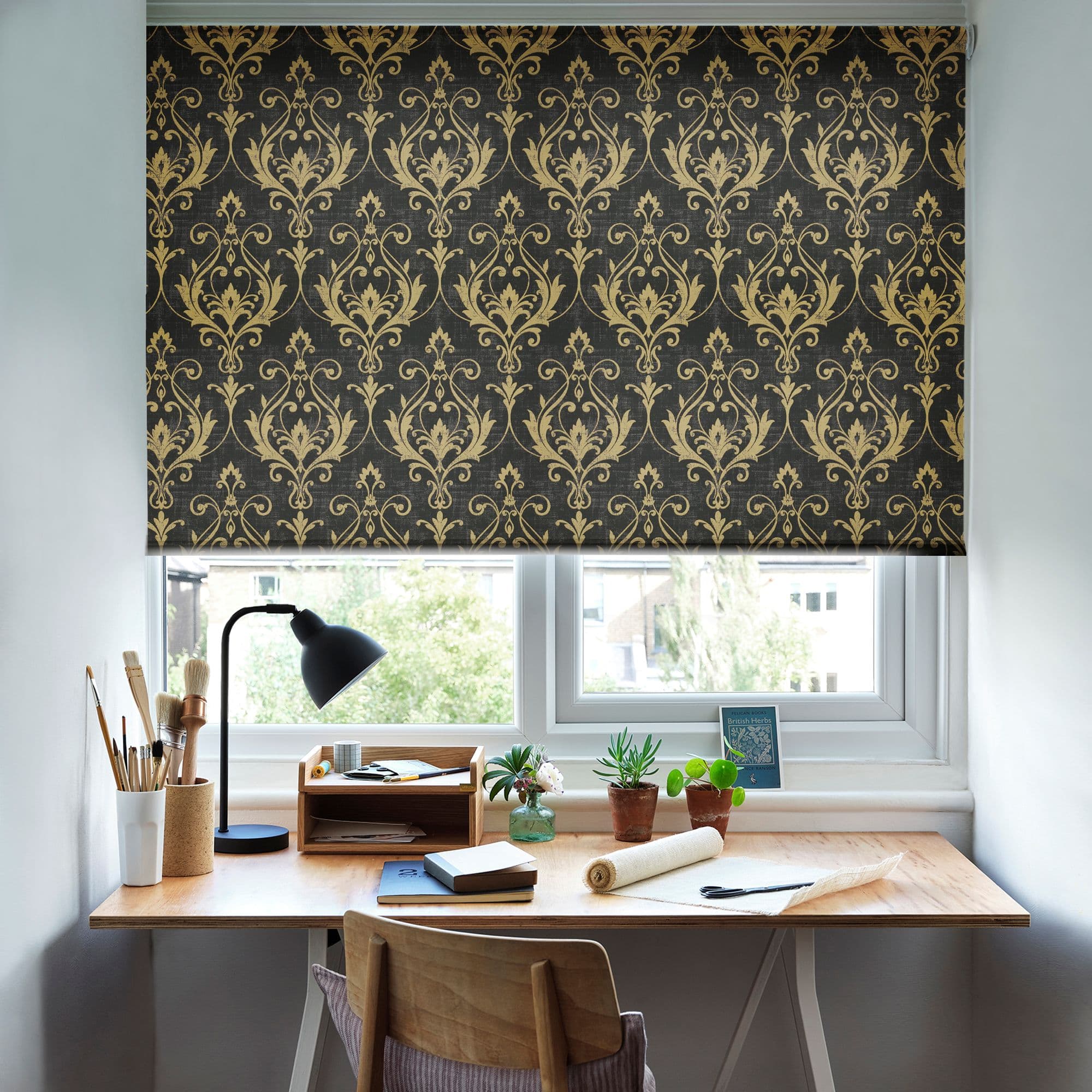 Roman blind black fabric with gold damask pattern lowered across upper window, filtering daylight; small home office desk beneath with lamp, art supplies, potted plants, and a view of trees and buildings.