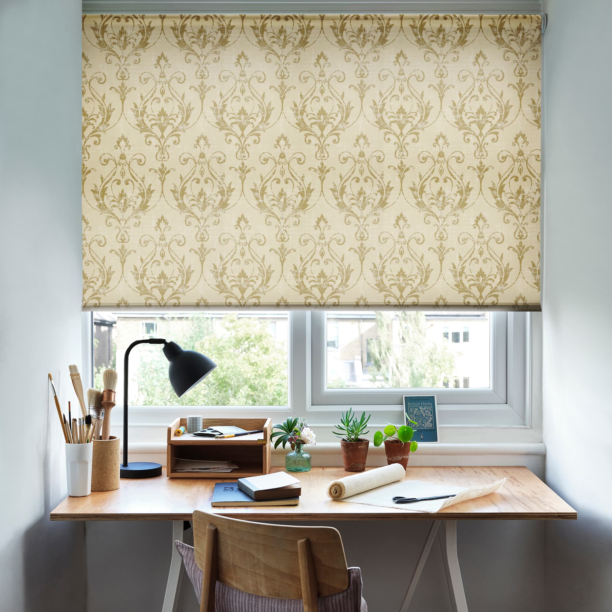 Beige patterned roller blind lowered over the window, damask motif filtering daylight; beneath, a wooden desk with lamp, brushes, notebooks and potted plants in a bright workspace with outdoor greenery.