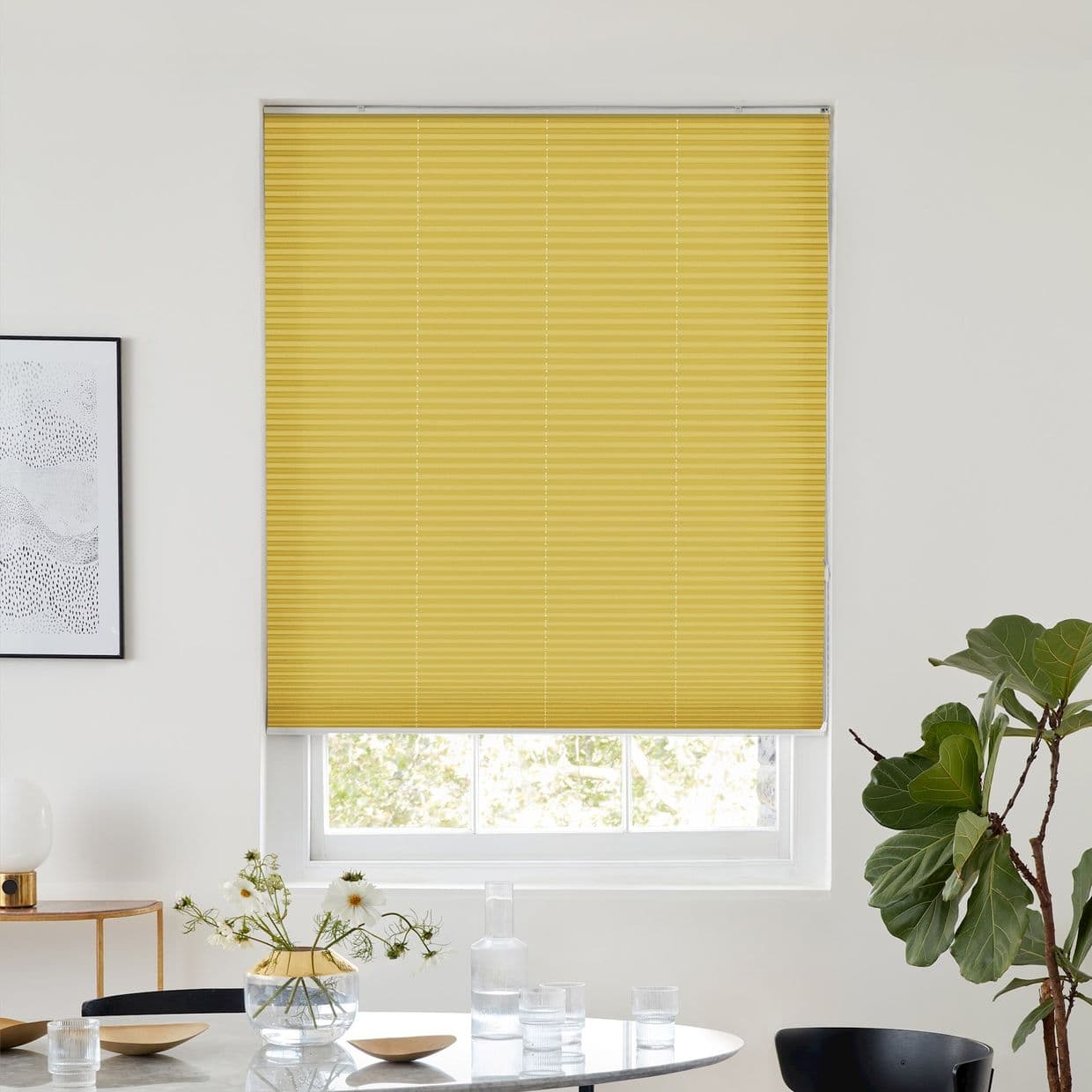 Yellow pleated blind lowered nearly to the sill, filtering light; set in a bright white dining room with a round table, glassware, framed art, and a potted fiddle-leaf fig.