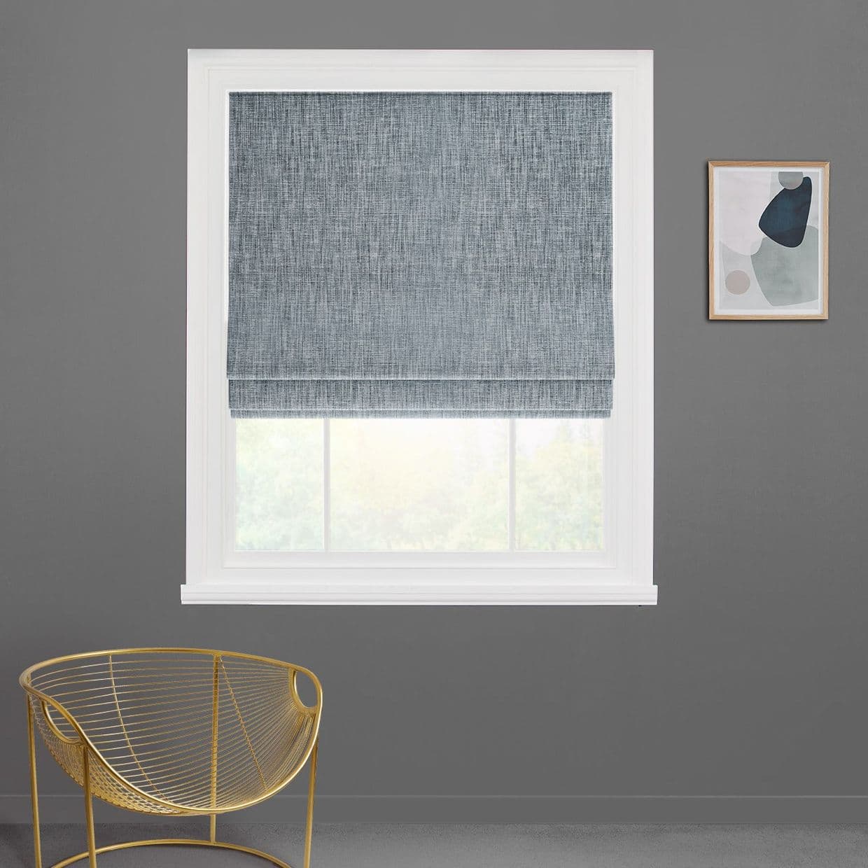 A textured gray-blue Roman shade, lowered mid-window with horizontal folds, filters daylight through a white-framed window in a gray-walled room with a gold wire chair and abstract art.