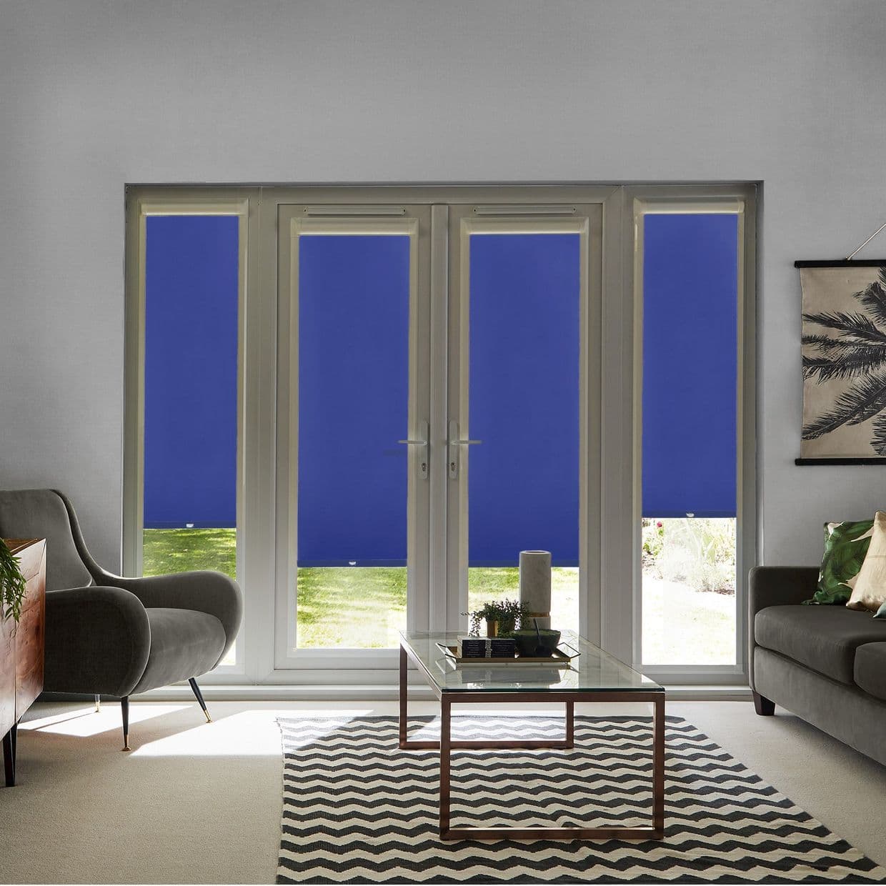 Blue roller blinds cover four-panel French doors and sidelights, lowered with bottom gaps showing lawn; they block and diffuse light in a living room with sofas, coffee table, chevron rug.