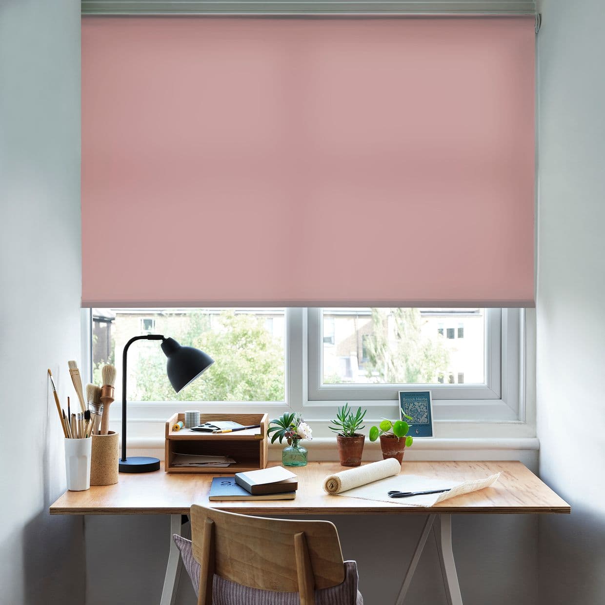 Pink roller blind, fully lowered with a slim lower gap, gently filtering daylight. Home workspace: wooden desk with black lamp, art brushes, notebooks and potted succulents beneath a double window.