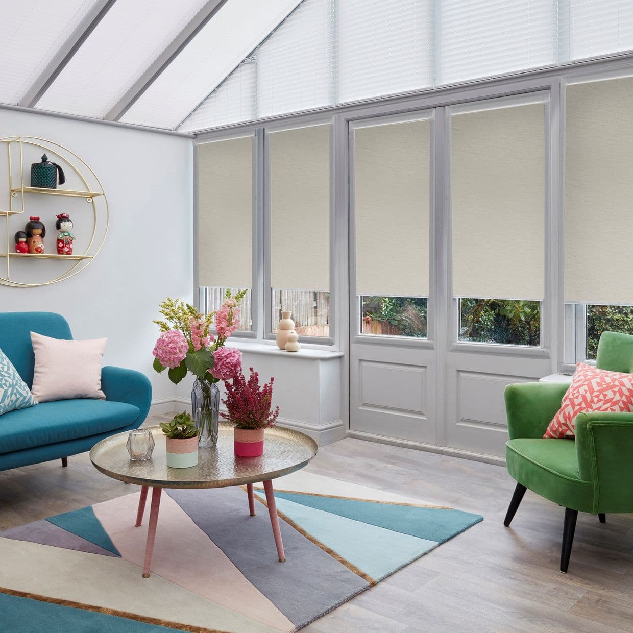 Beige roller blinds lowered across tall conservatory windows, filtering daylight mid-height; a bright living room with teal sofa, green armchair, round glass-top coffee table and pastel geometric rug.
