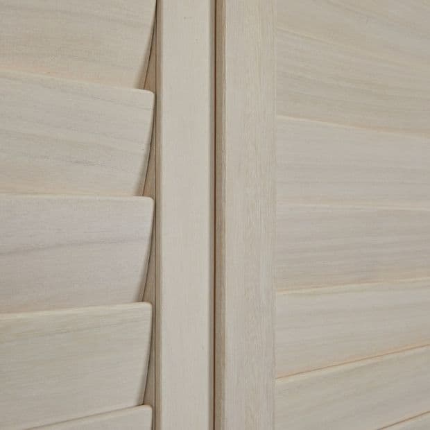 Pale wooden louvered shutters with a central vertical stile; left panel's slats are partially angled, right panel's slats closed, softly filtering interior light in a close-up of smooth wood grain.
