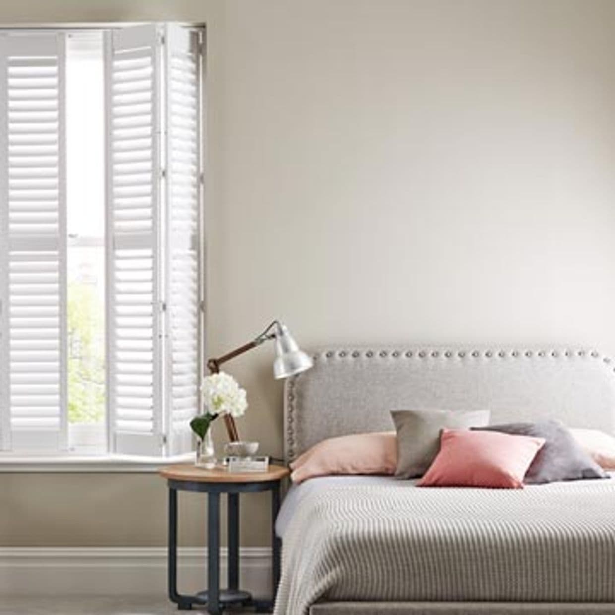 White wooden plantation shutters at left window, folded and partially open, filtering soft daylight into a neutral-toned bedroom with upholstered headboard, bedside table, lamp and cushions.