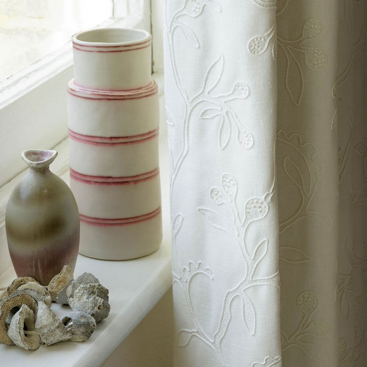 White embroidered curtain, hanging at the window's right edge with raised floral vine pattern; draped and filtering daylight; windowsill with stacked ceramic vases and shells.